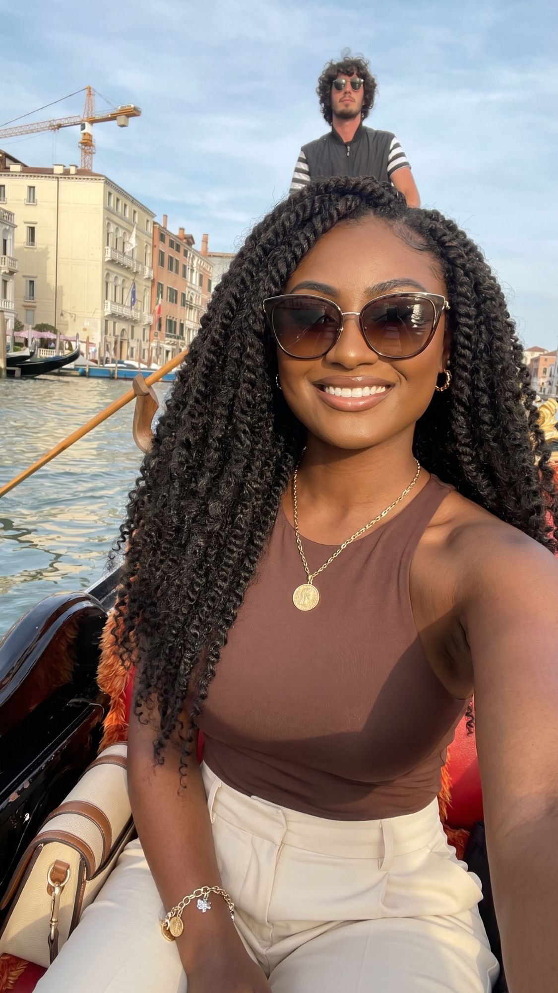 A woman with curly hair, wearing sunglasses, a brown sleeveless top, and cream pants, smiles while sitting in a gondola on a canal—an Italy bucket list moment—with buildings and a gondolier in the background.