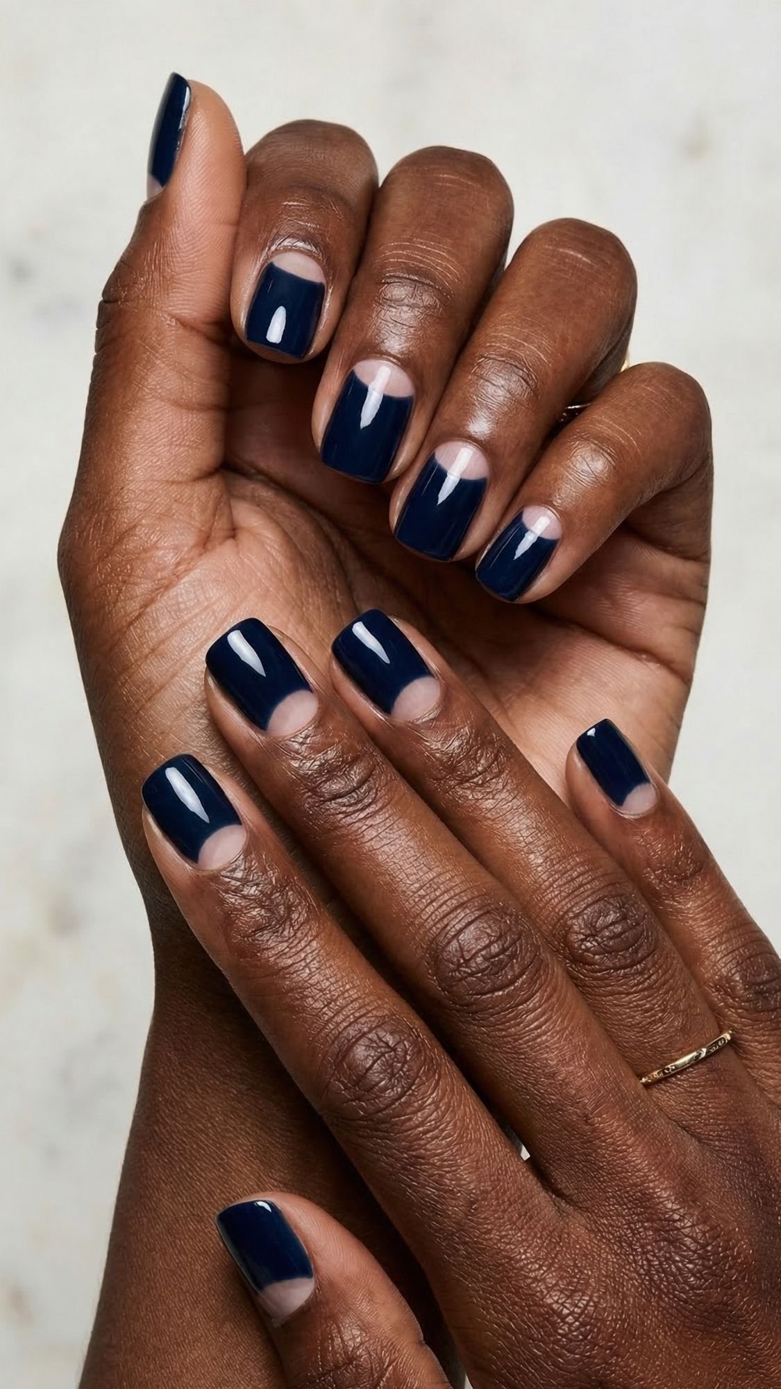 A close-up of hands with short, square-shaped nails painted in navy blue nail polish, leaving a crescent-shaped area near the cuticles unpainted for a modern half-moon manicure—an easy nail design perfect for simple nails at home.