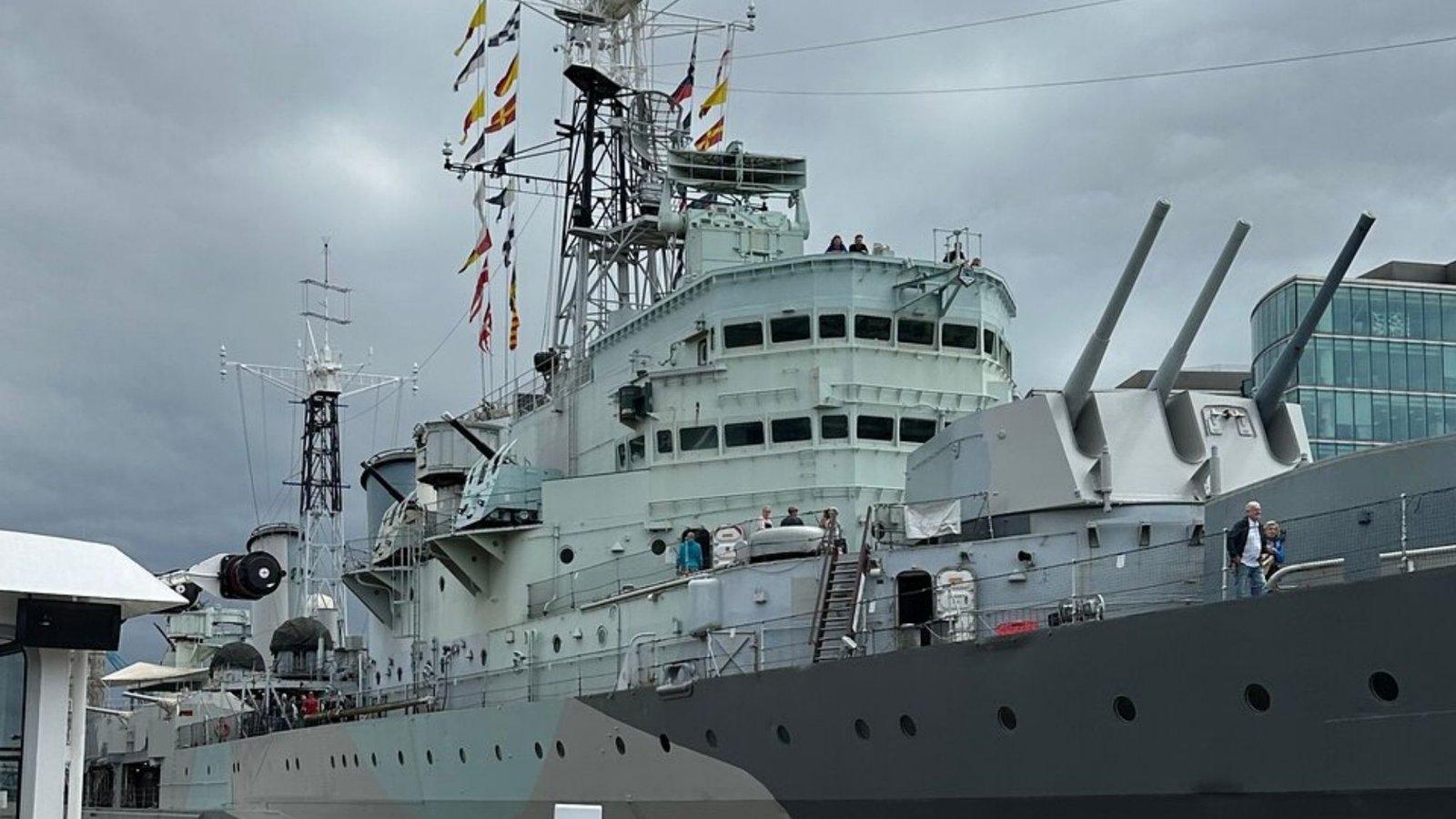 A large gray naval warship with multiple antennas, signal flags, and gun turrets docked at a port—a must-see for London travel. Several people explore the ship under a cloudy sky, making it perfect for your first trip to London.