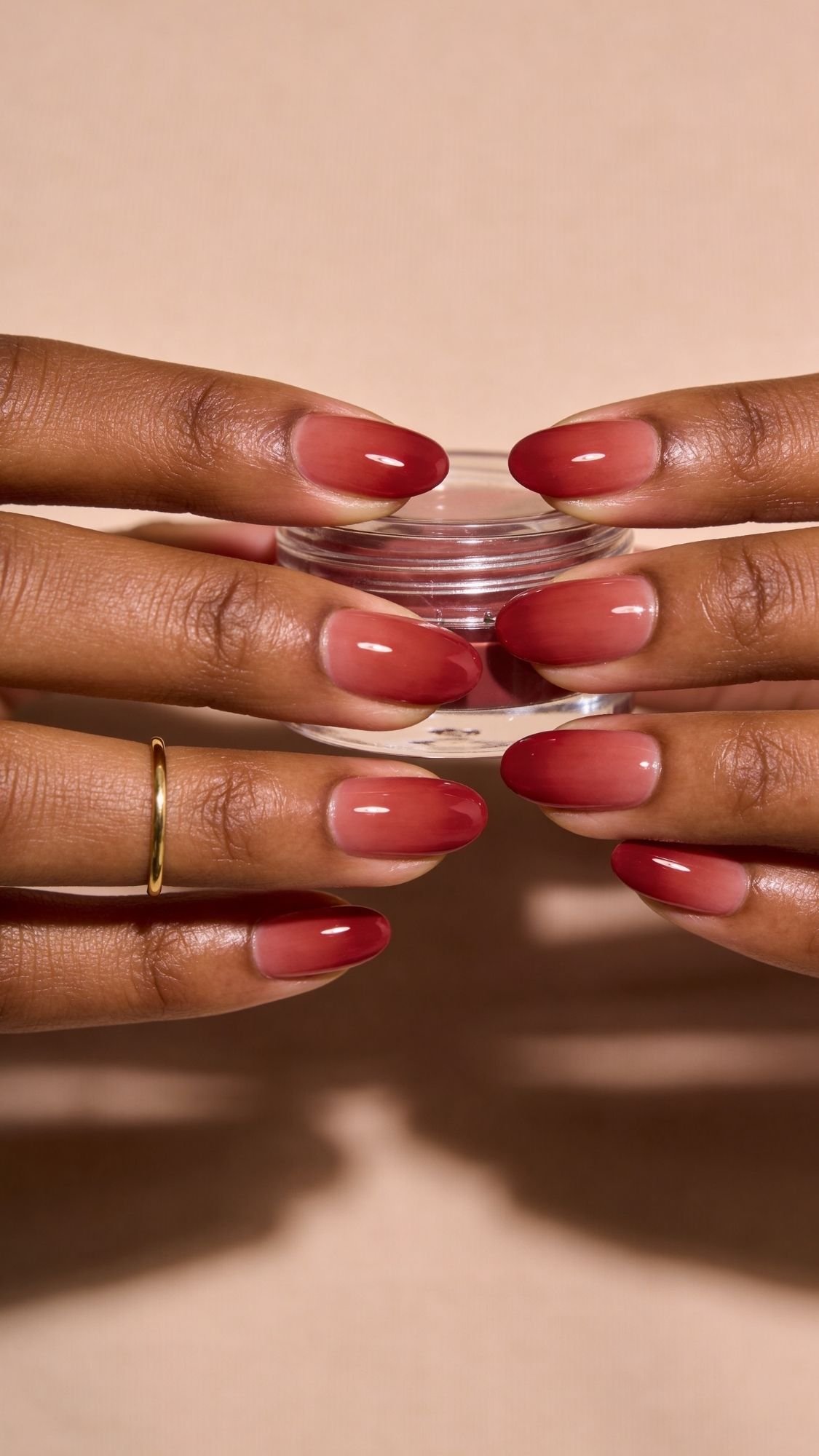 Two hands with almond-shaped nails painted in a red ombre design hold a small clear container. One hand has a simple gold band on the ring finger. This chic red nails look is perfect for every season and inspires fresh nail ideas.