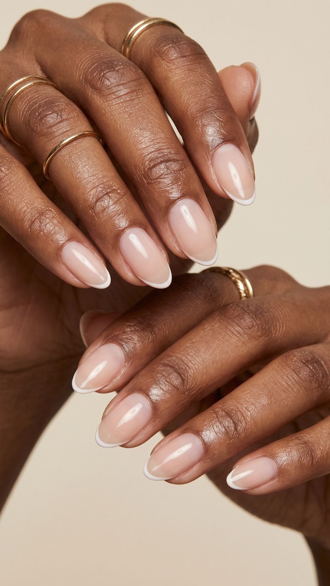 Close-up of hands with almond-shaped nails featuring a natural, glossy nude polish and thin white tips—an elegant choice for spring nails. Several thin gold rings adorn the fingers against a neutral background.