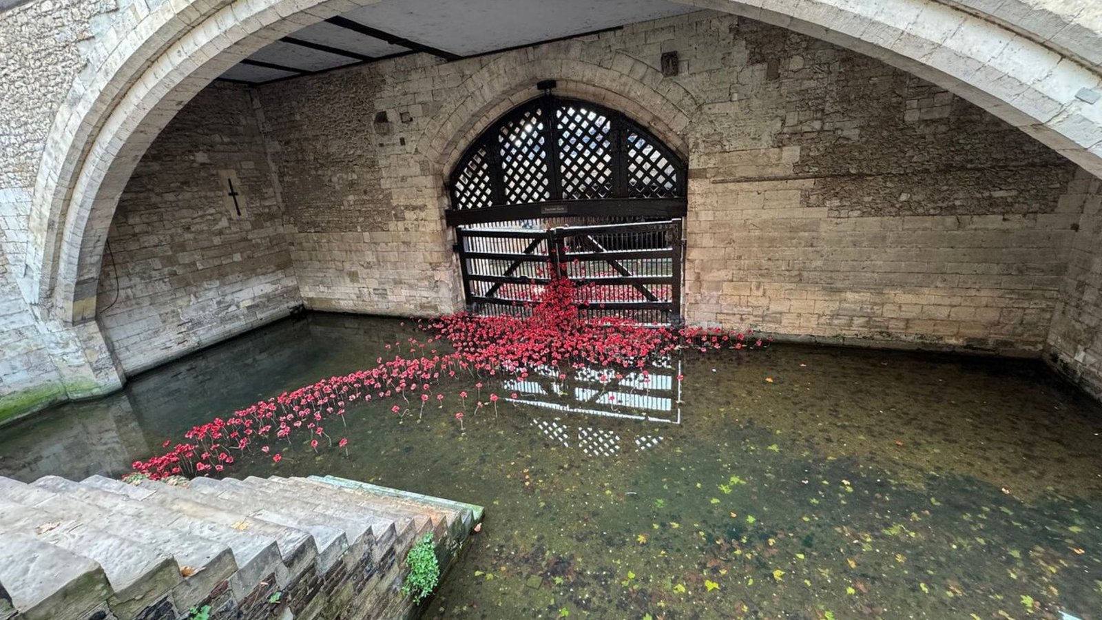 A historic stone arched gate over shallow water, with a cluster of red ceramic poppies at the base of the black iron gate, reflecting below—a striking sight among top London attractions for your first trip to London.