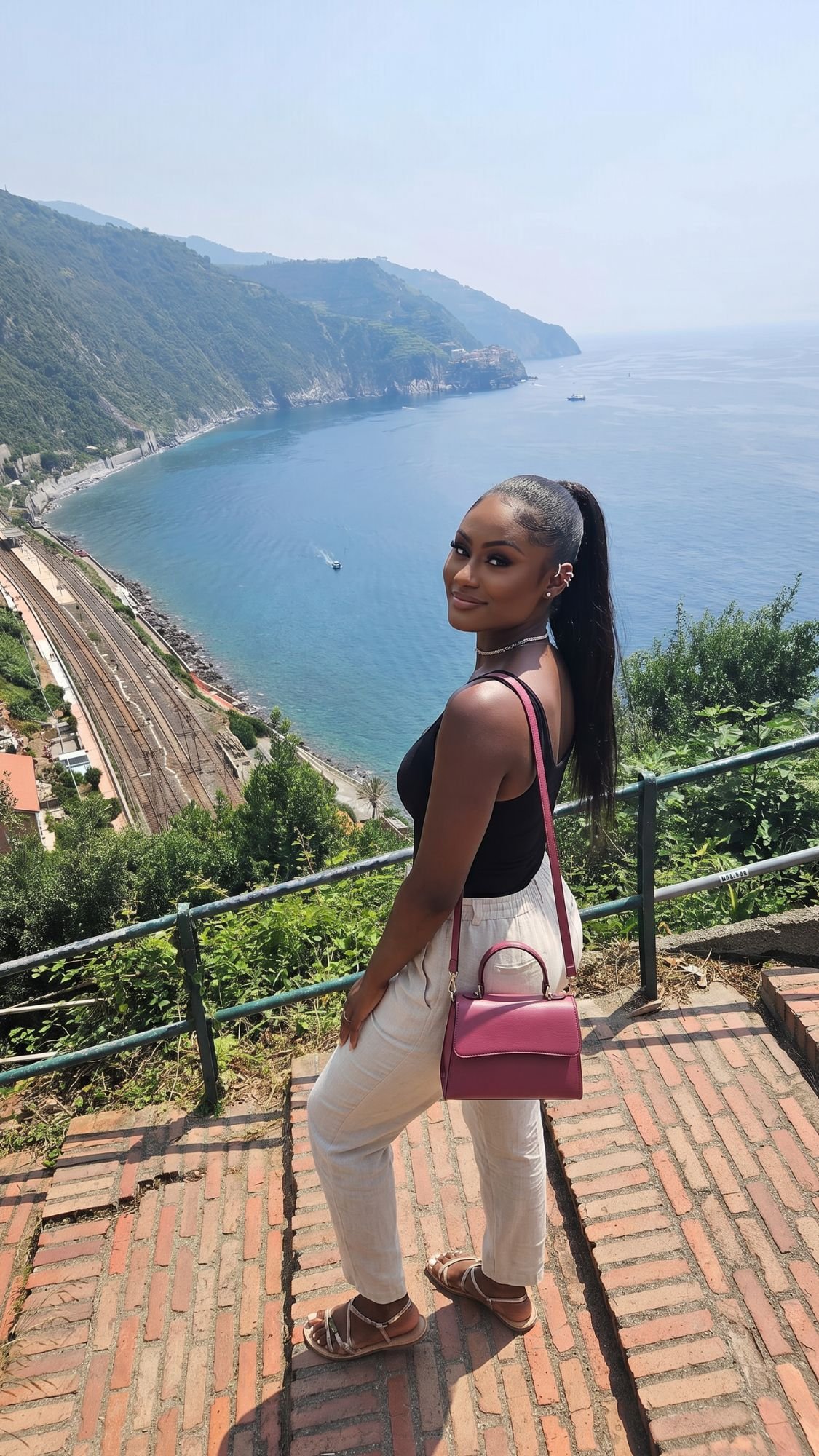 A woman stands on brick steps, smiling at the camera with a scenic coastal view, blue sea, green hills, and railway tracks in the background—capturing a perfect Italy bucket list moment on her first trip to Italy.