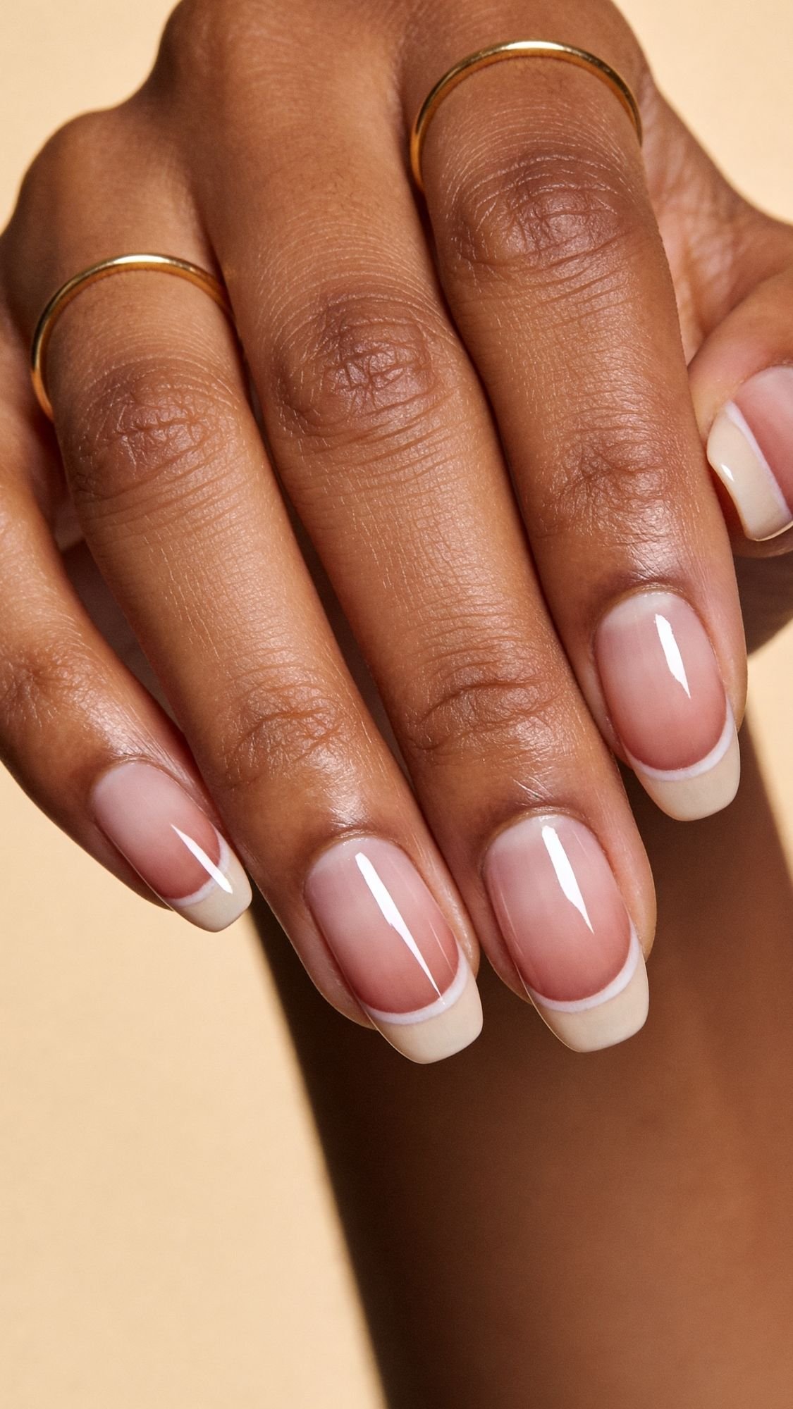 A close-up of a hand with a classic French manicure, perfect for spring nails, featuring natural-looking nails with white tips and a glossy finish. The hand is adorned with two thin gold rings against a soft beige background.