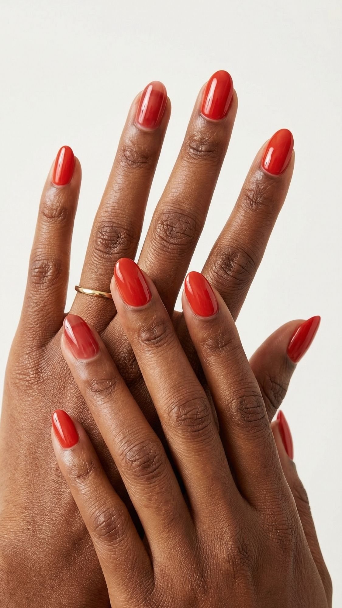 Two hands with dark skin tone display bright red nails, offering chic nail ideas for every season. One hand features a thin gold ring on the middle finger, set against a plain, light-colored background.