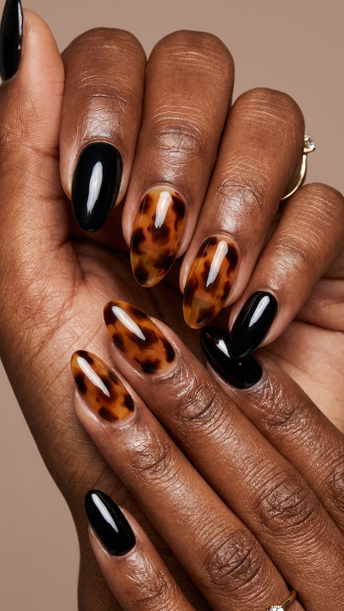 A close-up of hands with manicured nails featuring a mix of glossy black polish and amber tortoiseshell designs on an almond shape, against a neutral background—a chic example of easy nail designs you can try for stylish nails at home.