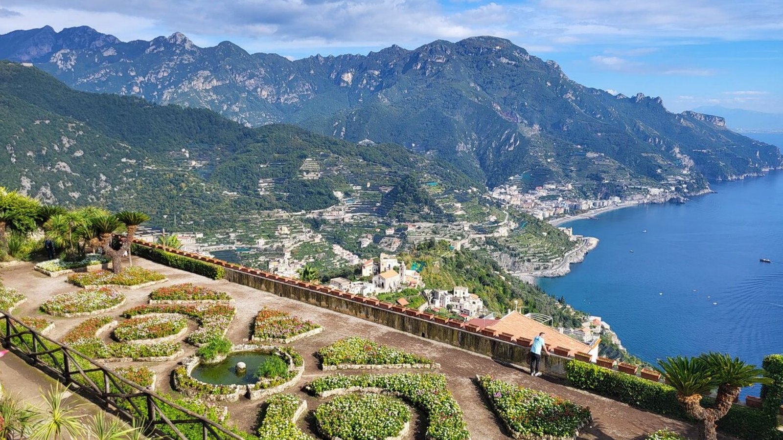 A terraced garden with flowerbeds and a small round pond overlooks a scenic coastline, inviting you to add this view to your Italy bucket list—where steep green mountains, blue sea, and charming villages await under a partly cloudy sky.