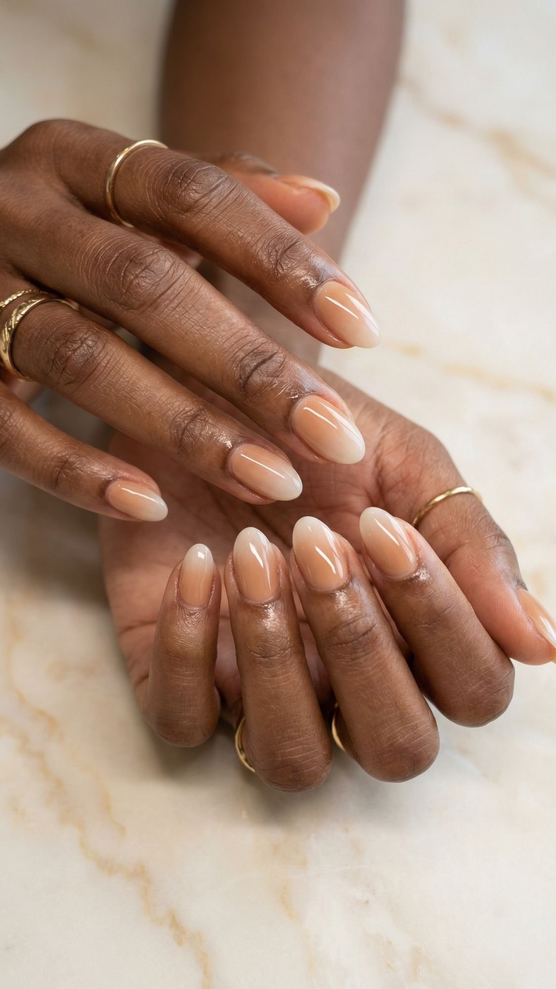 Close-up of hands with almond-shaped nails painted in a glossy nude gradient, perfect for summer nails. The person is wearing thin gold rings on several fingers, set against a light marble background.