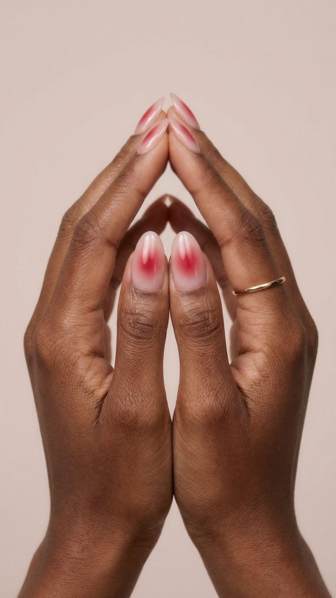 Two hands with dark skin are held together, fingertips touching to form a flame shape. The almond-shaped nails feature pink gradient seasonal nails, and a thin gold ring adorns one finger. The background is light beige.