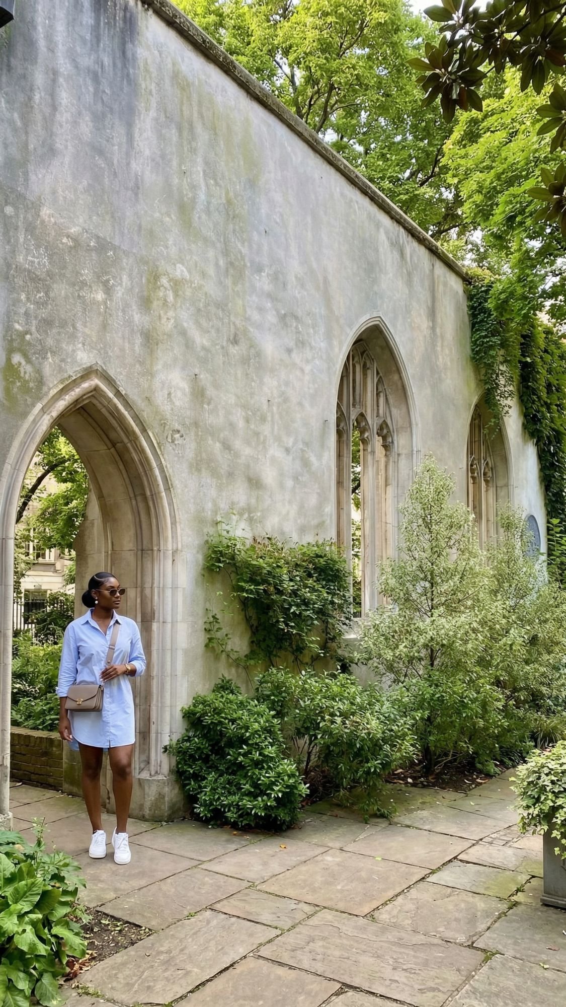 A woman in a light blue dress stands by arched stone windows of a greenery-covered ruin, wearing sunglasses and white sneakers—discover hidden gems like this on your first trip to London, exploring unique London attractions.