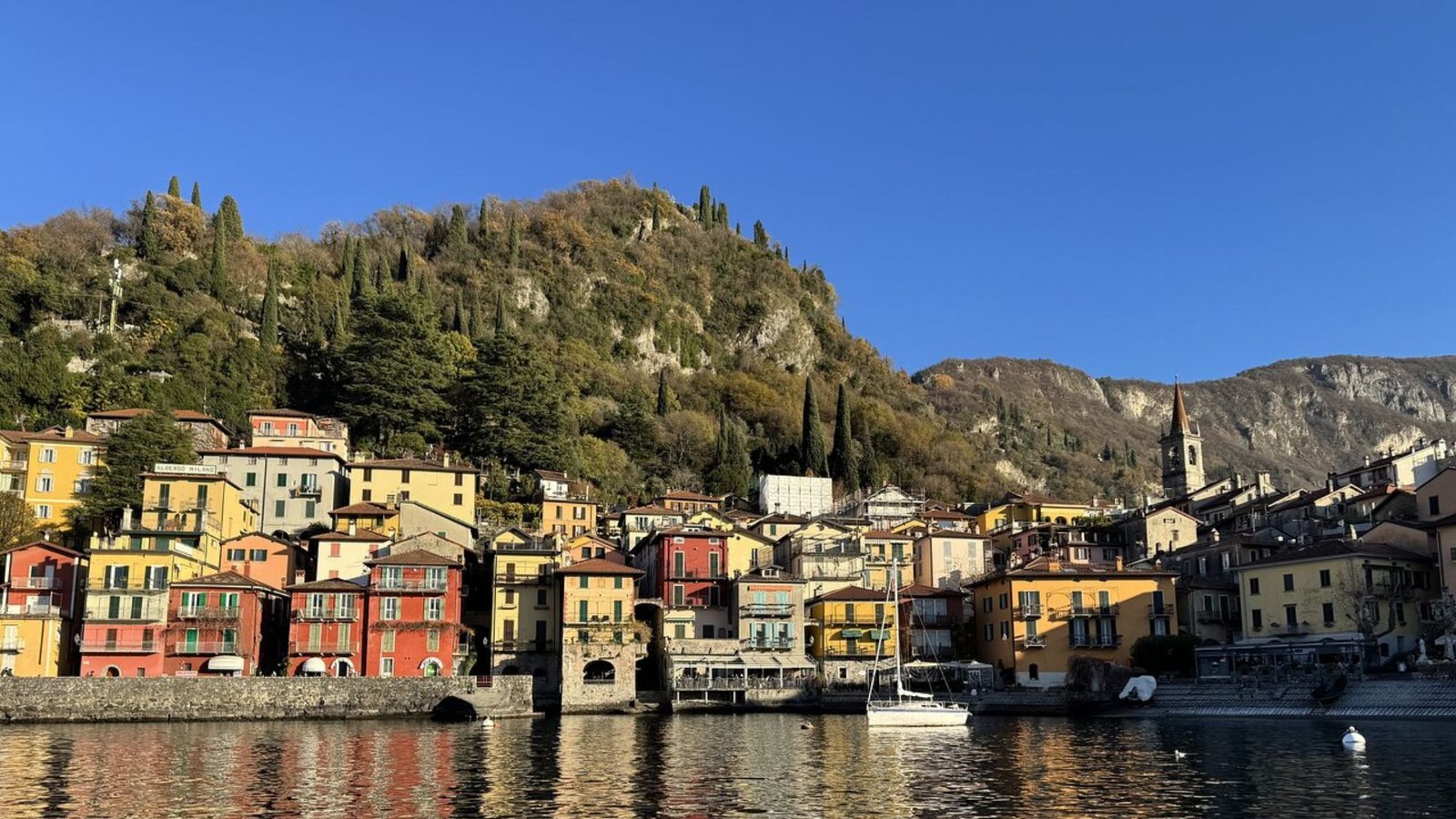 Colorful buildings line the waterfront in a picturesque village at the base of a tree-covered hillside—an unforgettable sight for anyone’s Italy bucket list, with clear blue skies above and shimmering reflections in the calm water below.
