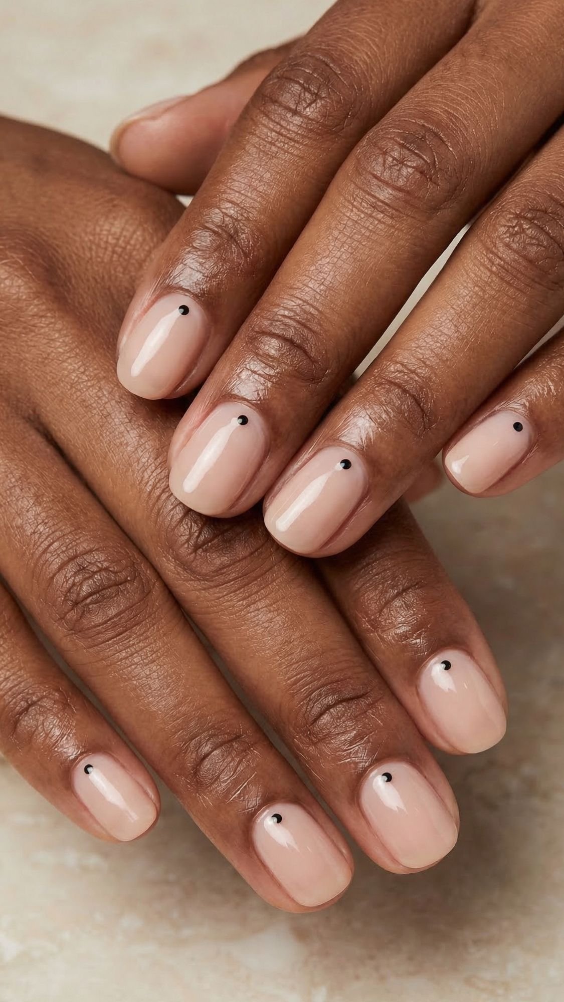 A close-up of hands with short, nude-colored polished nails, each featuring a small black dot near the base—perfect for easy nail designs you can try on your simple nails at home.