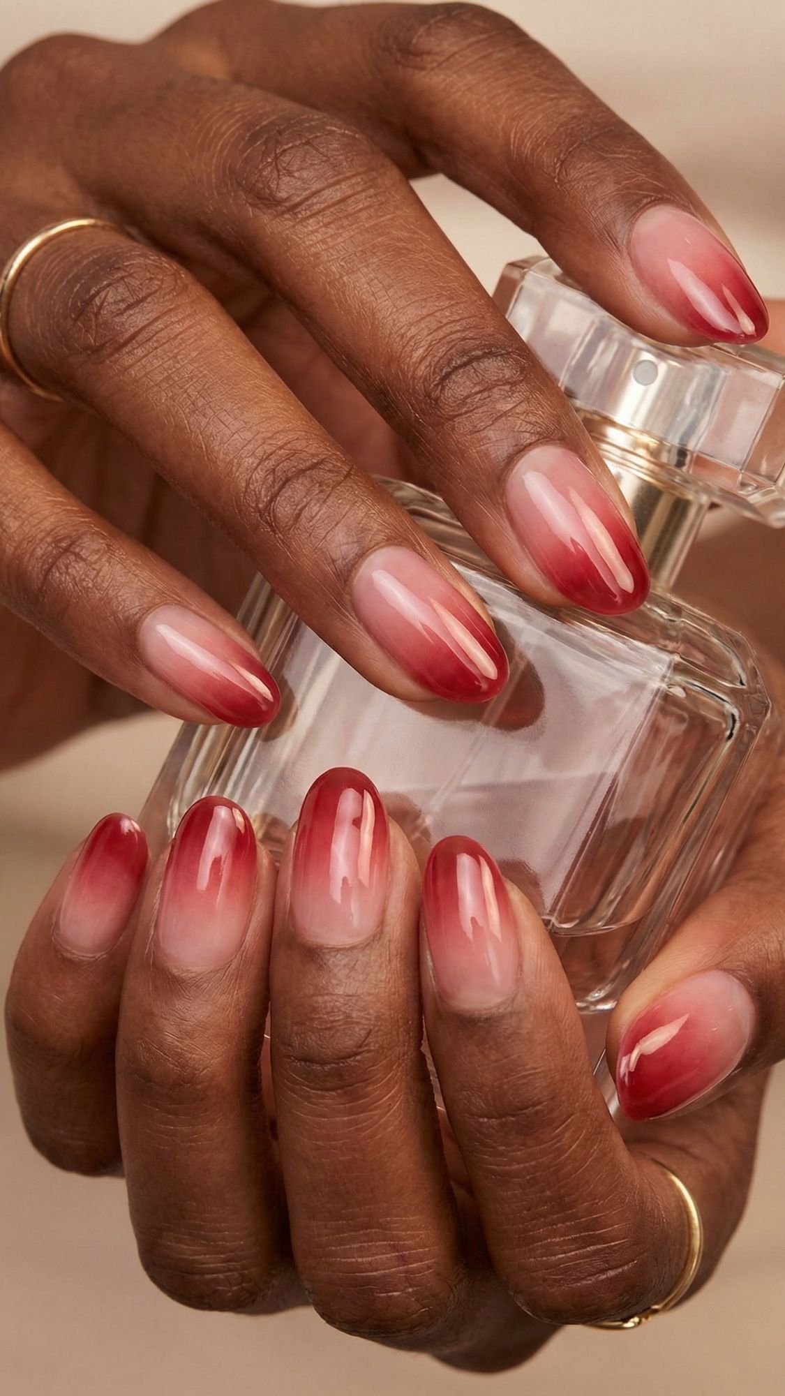 A close-up of hands with red ombre nails holding a glass perfume bottle. The almond-shaped manicure is one of the chicest red nail designs, and thin gold rings adorn two fingers.