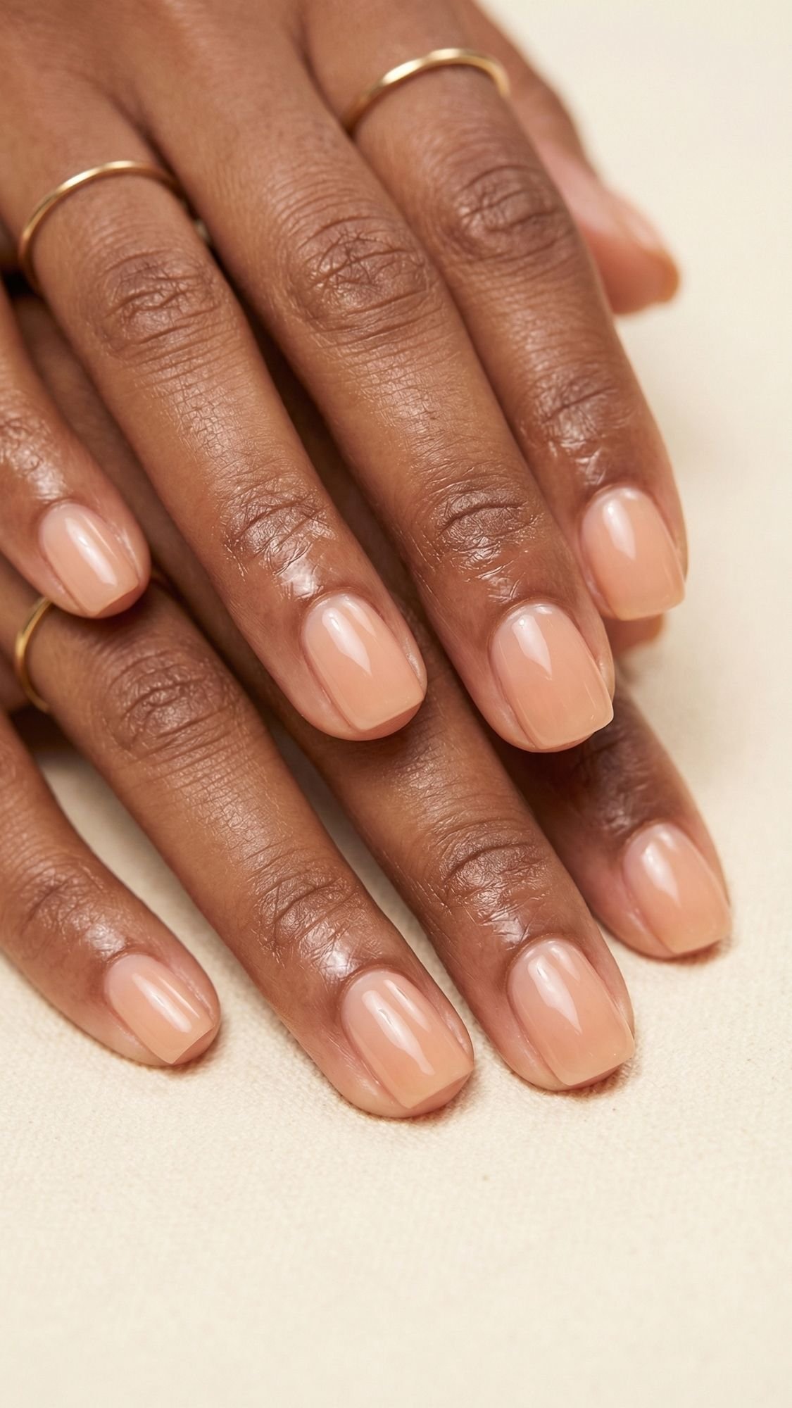 Close-up of two hands with neatly manicured nails painted in a glossy nude polish—perfect for summer nails. The person wears thin gold rings, and the background is a light neutral color, offering chic nude nail ideas.