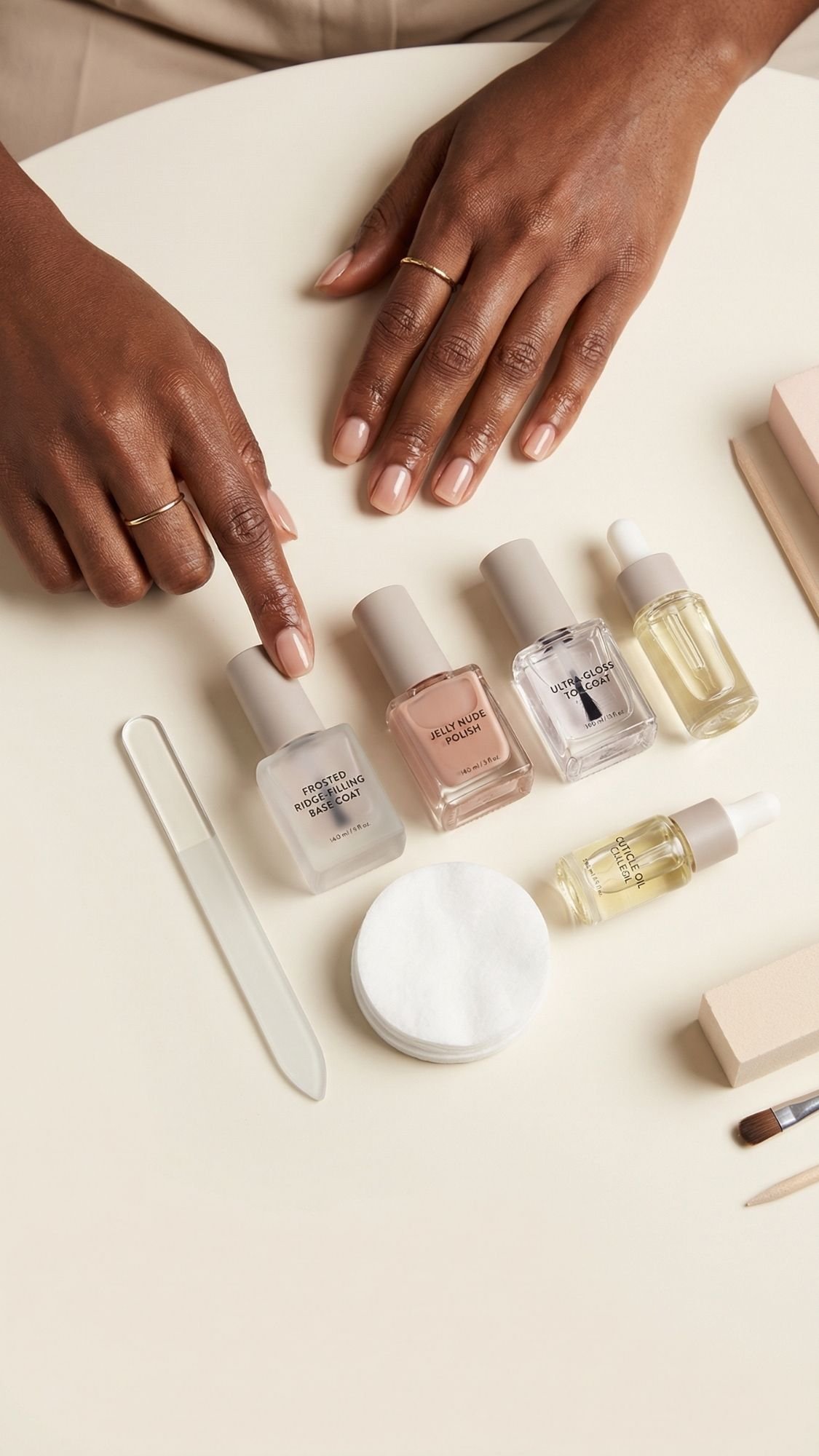 A person with neatly manicured nails points to a bottle of nail polish on a table filled with summer nails essentials, including four polish bottles, two dropper bottles, a nail file, cotton pads, and other manicure tools.