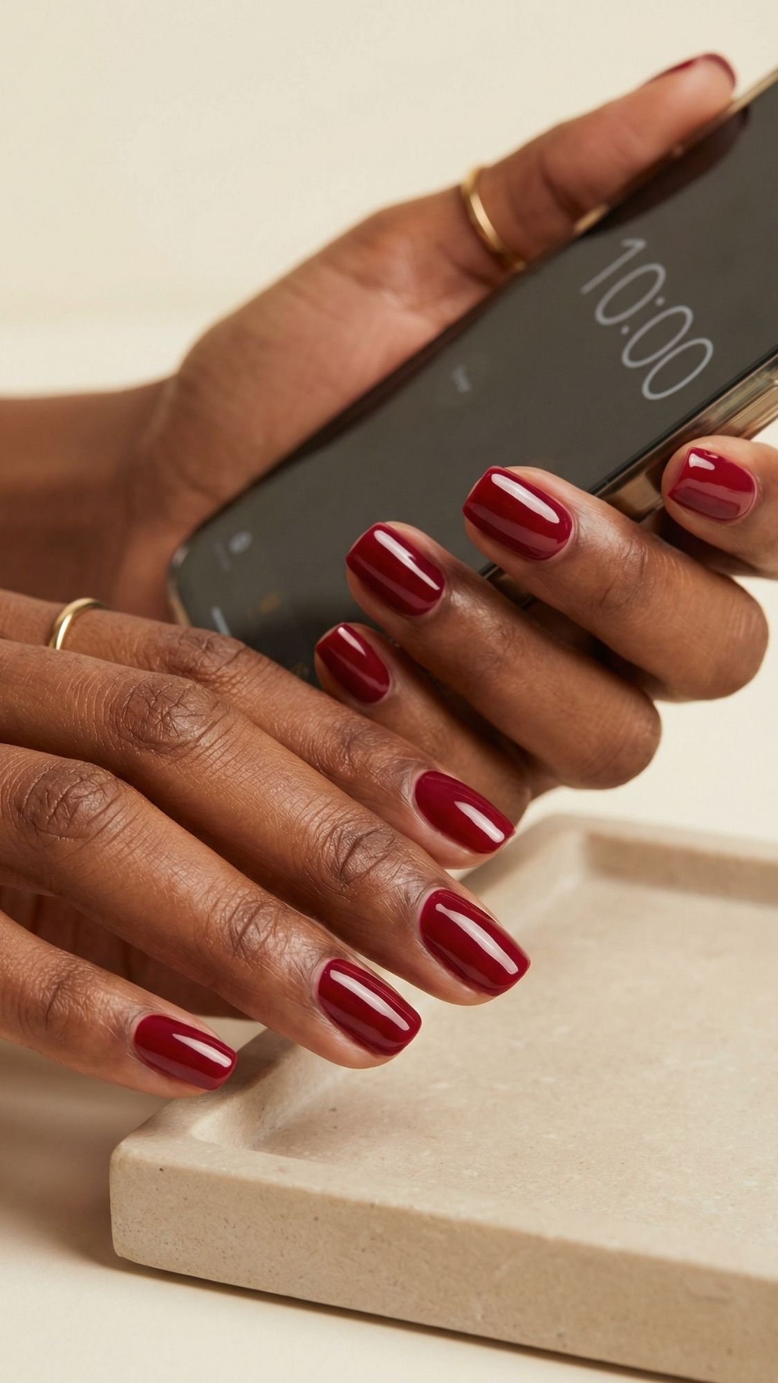 A person with neatly manicured Red nails holds a smartphone showing the time 10:00, with a beige tray and surface in the background—perfect inspiration for red nails ideas or seasonal nail designs.