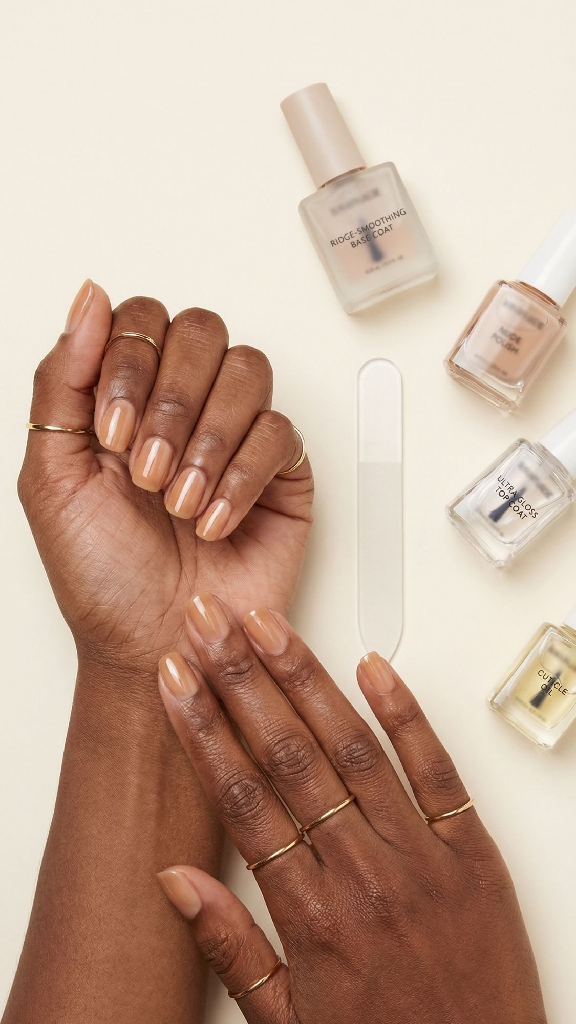 A person with neatly manicured, nude-colored nails rests their hands on a beige surface. Several rings adorn their fingers, showcasing chic nude nail ideas. Nail polish bottles and a glass nail file are arranged beside the hands.
