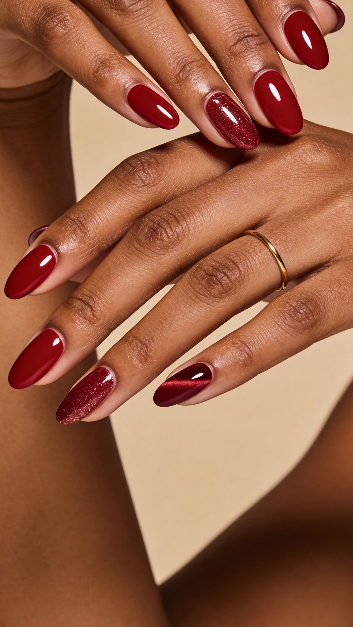 Close-up of hands with long, almond-shaped red nails—one on each hand features red glitter polish for a sparkling accent. The person wears a simple gold ring, making this look perfect for seasonal nails or fresh nail ideas. Beige background adds warmth.