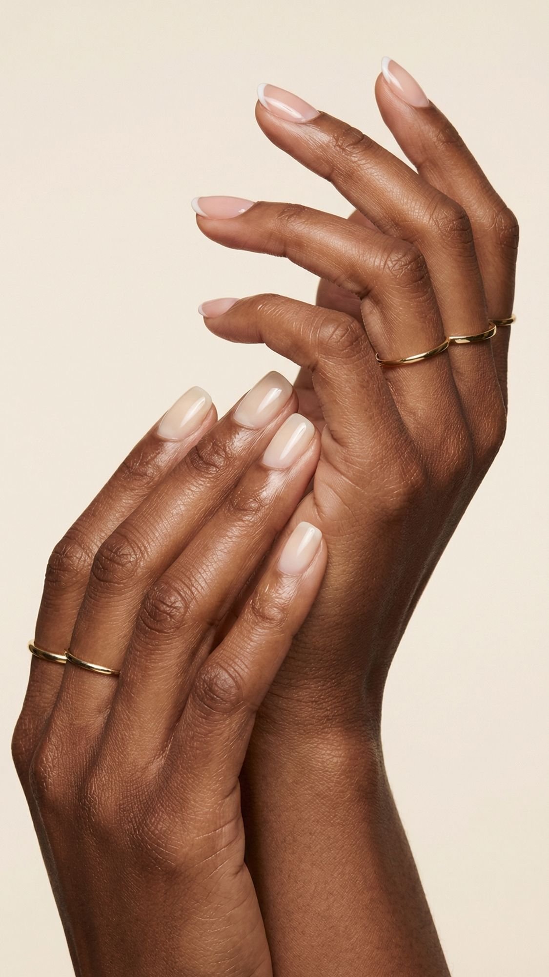 Two hands with dark skin are gracefully posed against a light background. The neatly manicured nails feature a natural, nude polish—perfect for summer nails—and each hand is adorned with simple gold rings on the index and middle fingers.