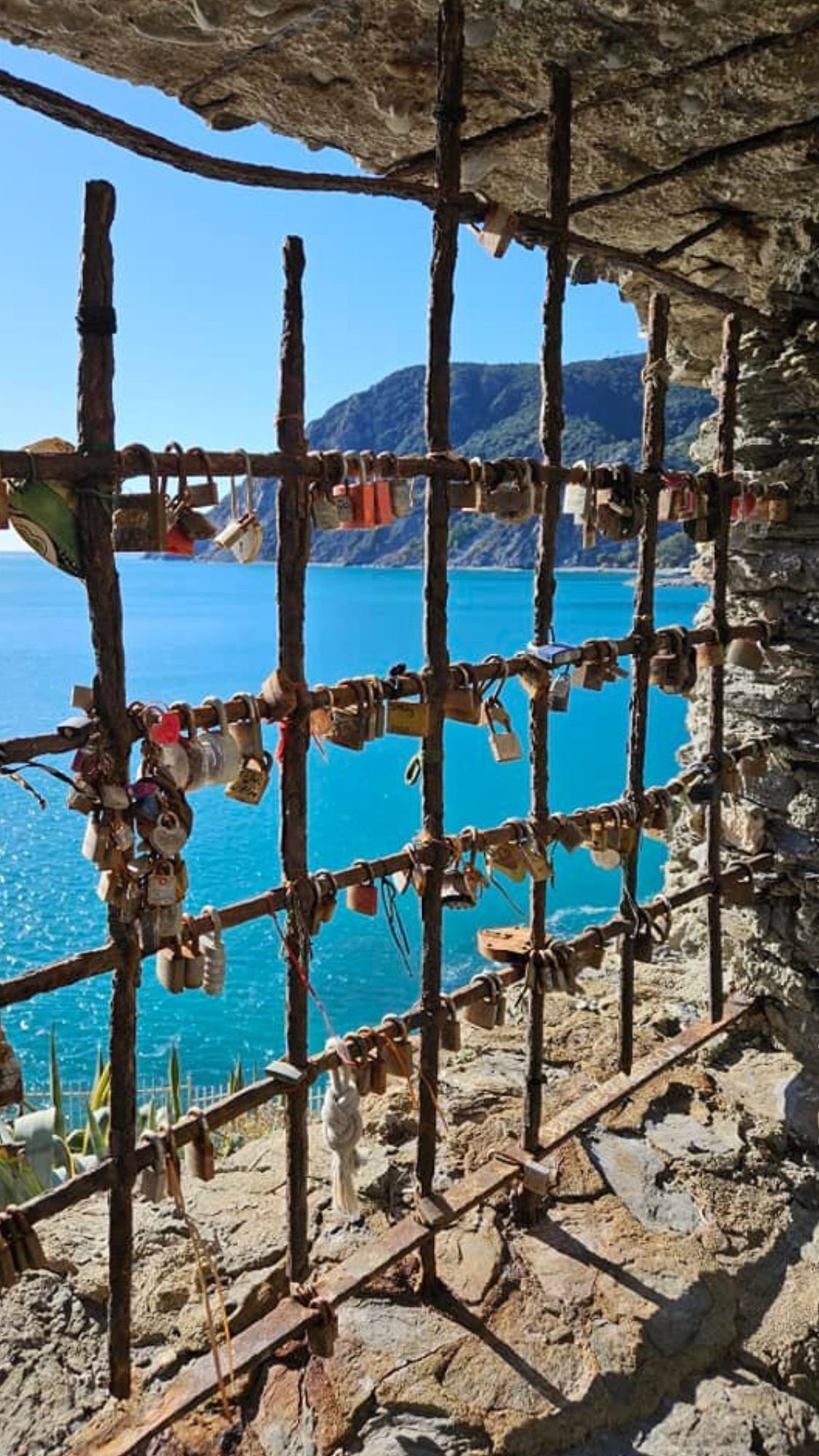 Rusty iron bars covered with numerous padlocks overlook a bright blue sea and distant rocky cliffs through a stone window, sunlight illuminating a scene straight from any Italy bucket list.
