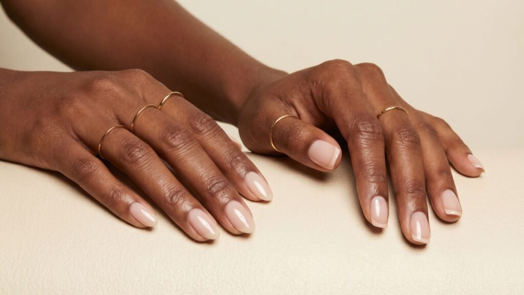 Two hands with neatly manicured, nude-colored nails—perfect for summer nails—inspire fresh style. Each hand is adorned with several thin gold rings, beautifully highlighting the medium to dark brown skin tone.