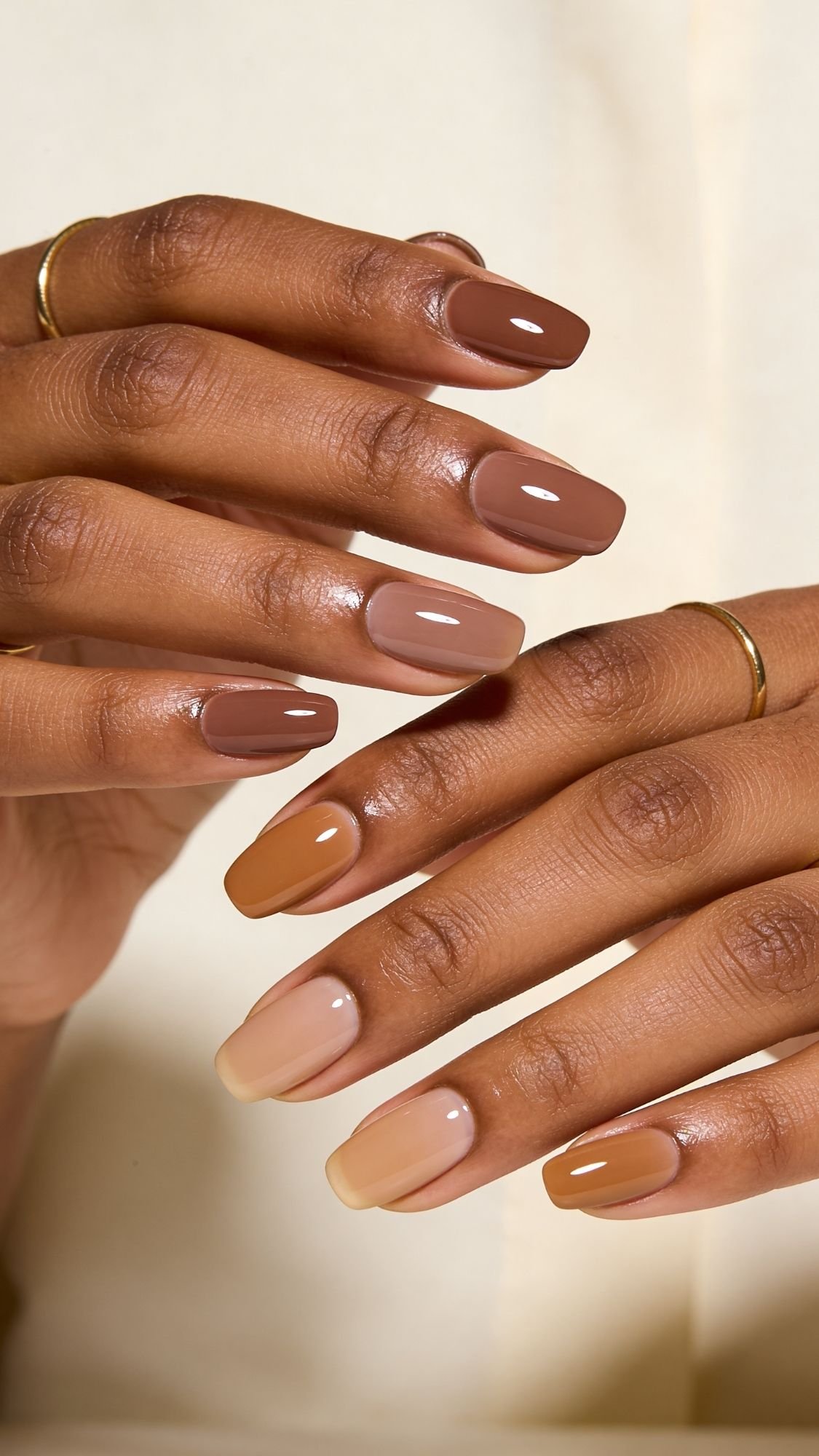 A close-up of hands with manicured nails painted in varying shades of nude and brown, showing a gradient effect—perfect for spring nails. The person wears thin gold rings on several fingers against a light background.