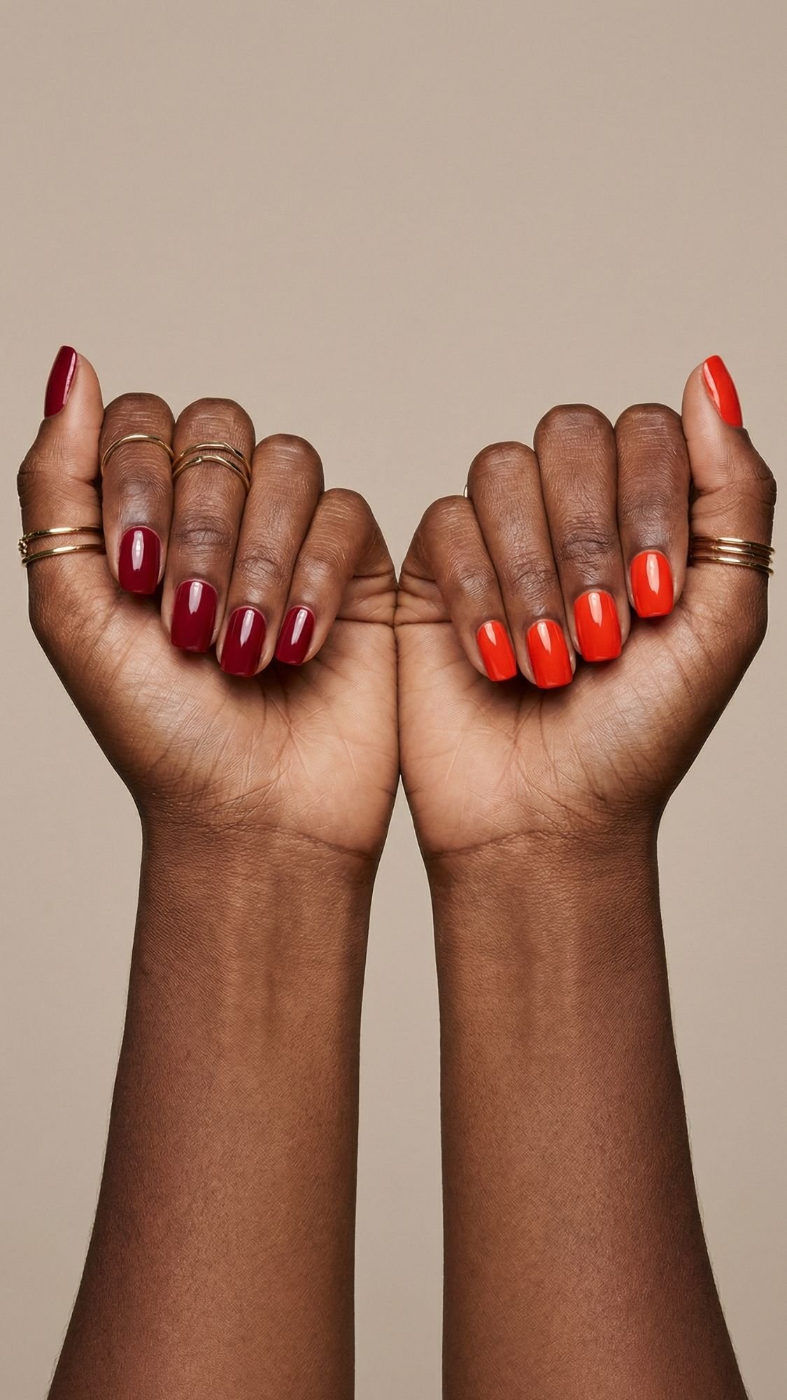 Two hands with medium-dark skin tone are held up against a beige background, showing off chic red nails in alternating shades of deep red, maroon, and bright orange—an inspiring nail idea for every season. Thin gold rings add a touch of elegance.