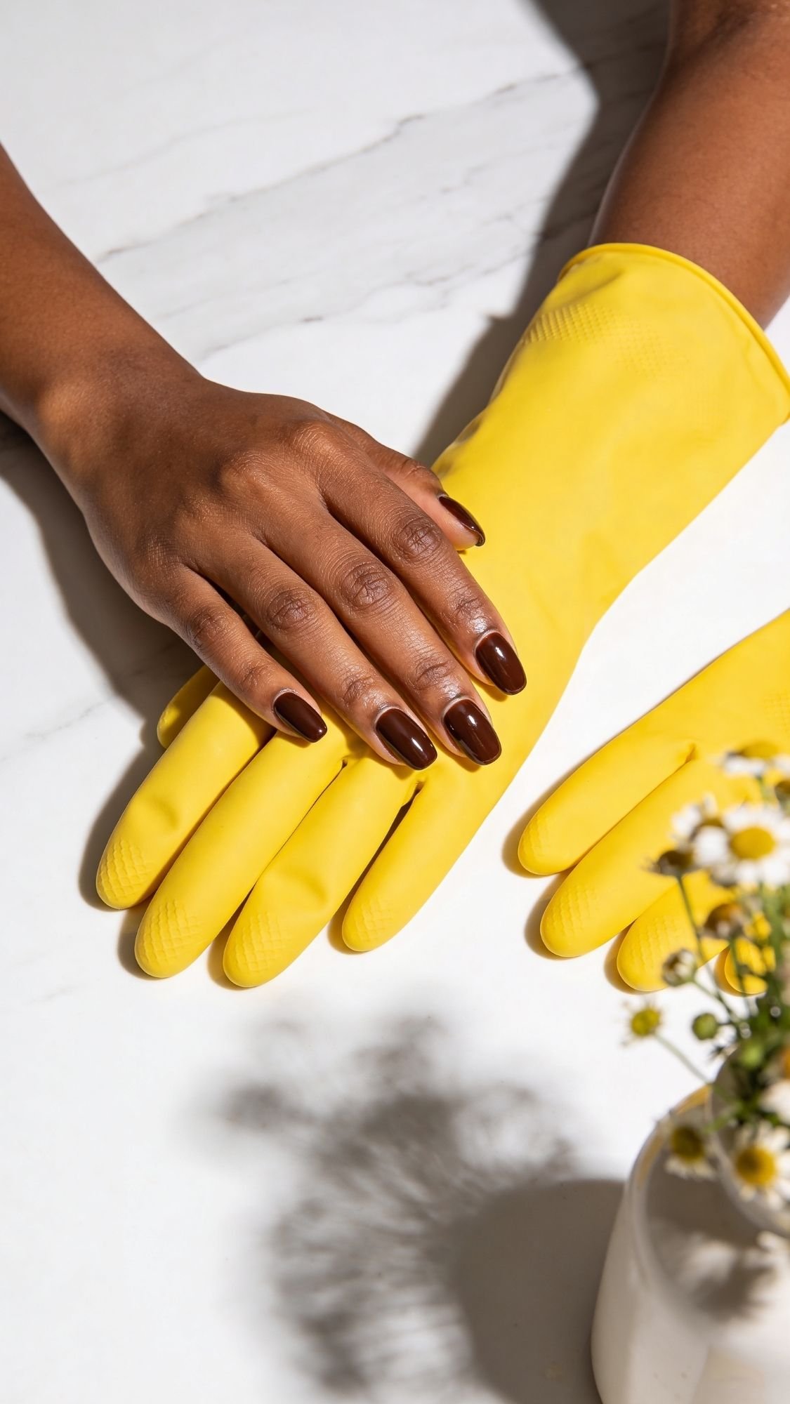 A person with dark polished nails, showcasing easy nail designs, rests their hand on bright yellow rubber gloves on a white surface, with a vase of small white and yellow flowers in the corner.