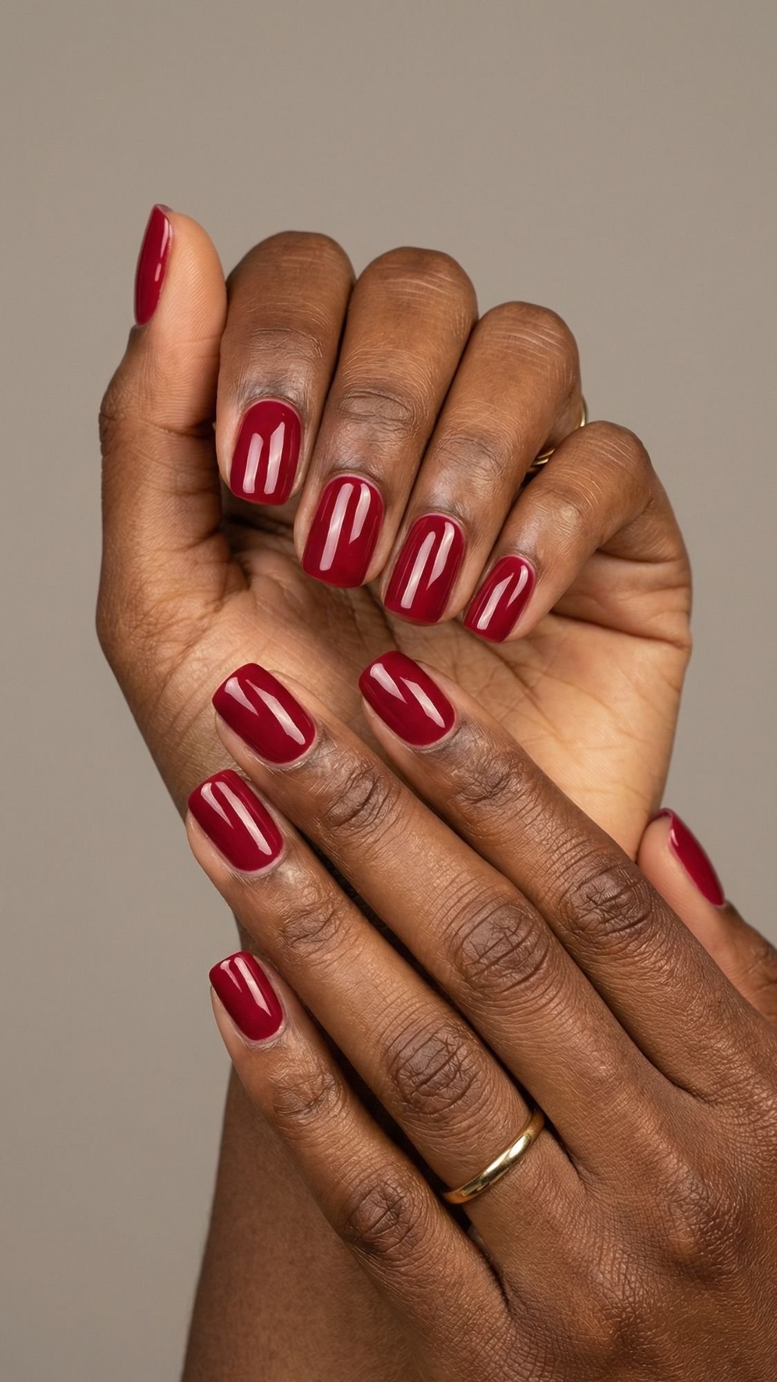 A close-up of two hands with dark skin, featuring neatly manicured red nails painted in a glossy deep hue. One hand gently rests on the other against a neutral background, making this a perfect inspiration for seasonal nails.