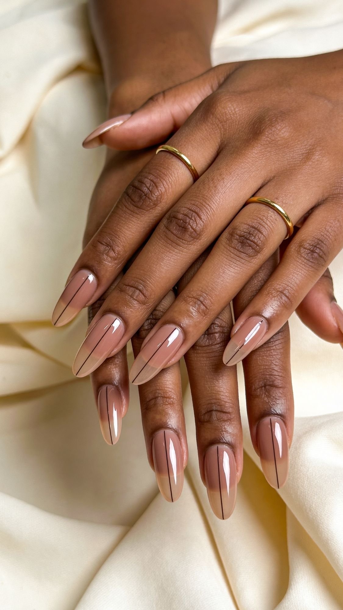 Two hands with almond-shaped nails featuring a nude base and a single dark vertical line on each nail—a chic choice for spring nails. The hands rest on cream-colored fabric, and the fingers are adorned with thin gold rings.