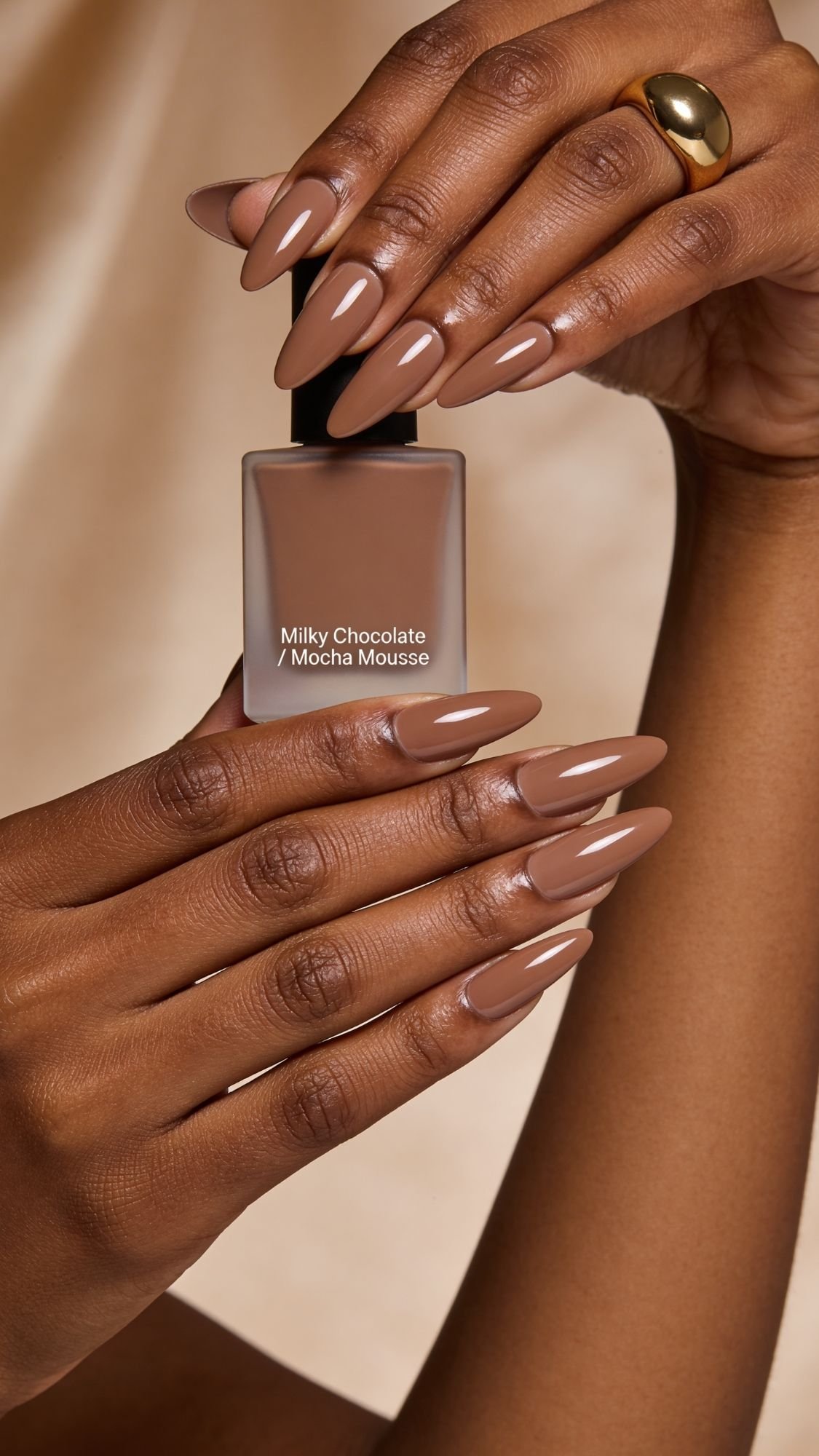 A hand with long, almond-shaped nails painted in a glossy mocha brown holds a matching bottle of nail polish labeled Milky Chocolate / Mocha Mousse against a beige background—perfect for simple nails or easy nail designs you can recreate at home.