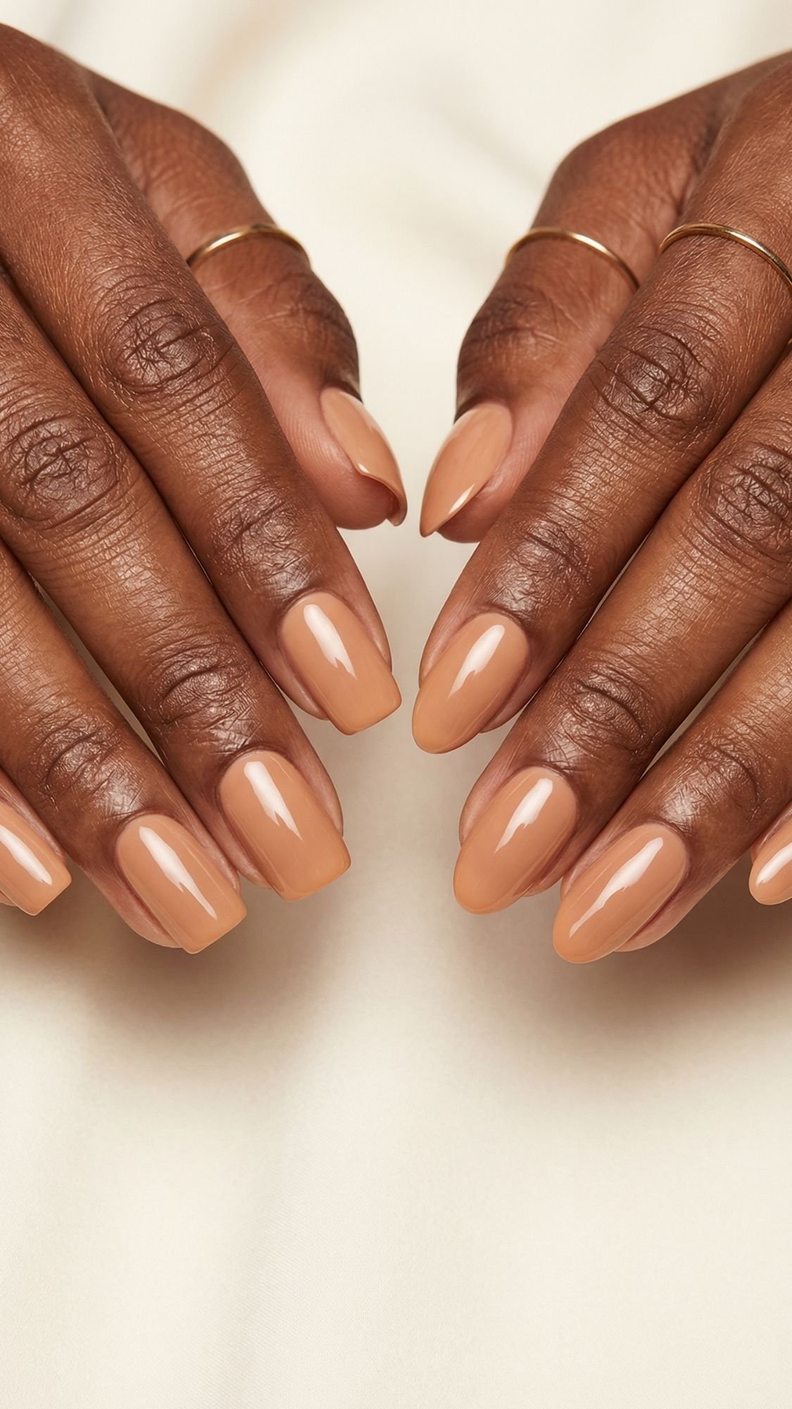 Close-up of hands with medium-brown skin, wearing thin gold rings, showing neatly manicured nails painted with glossy, nude beige polish—perfect inspiration for spring nails or anyone seeking chic nude nail ideas against a soft light background.