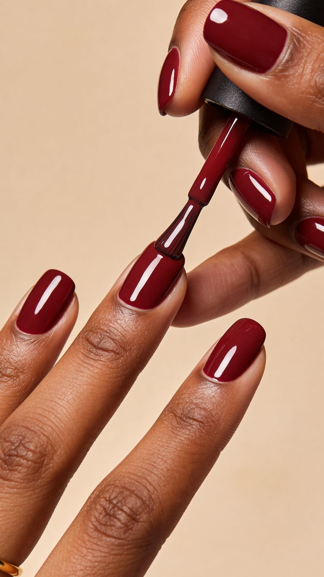 A close-up of a hand applying deep red nail polish to neatly manicured nails against a beige background—perfect Red nails inspiration for every season and fresh nail ideas.