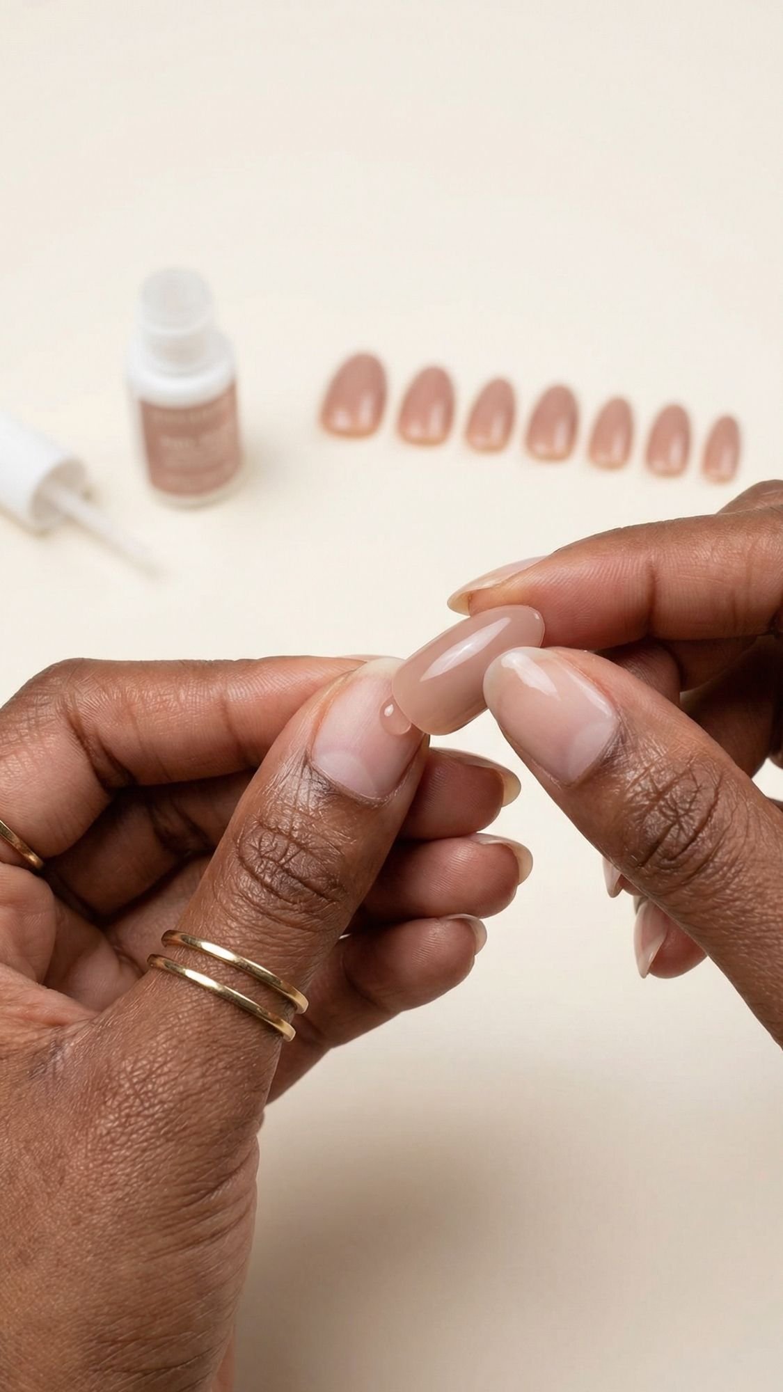 Close-up of hands with neutral nail polish applying a nude-colored press-on nail—perfect for Summer Nails. In the background, more press-on nails and a small bottle of nail glue rest on a light surface.