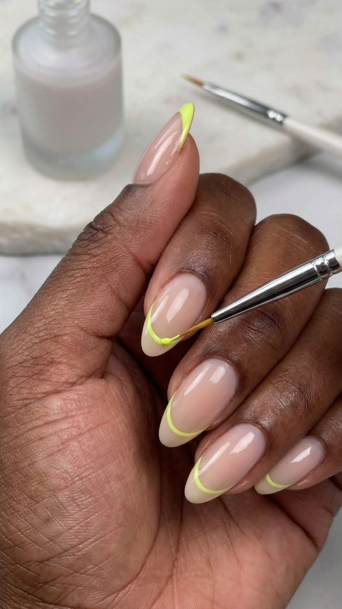 A hand with almond-shaped simple nails is being painted with a thin neon yellow French tip using a small brush. Perfect for easy nail designs and creating stylish nails at home, with a nude polish bottle and brush resting in the background.