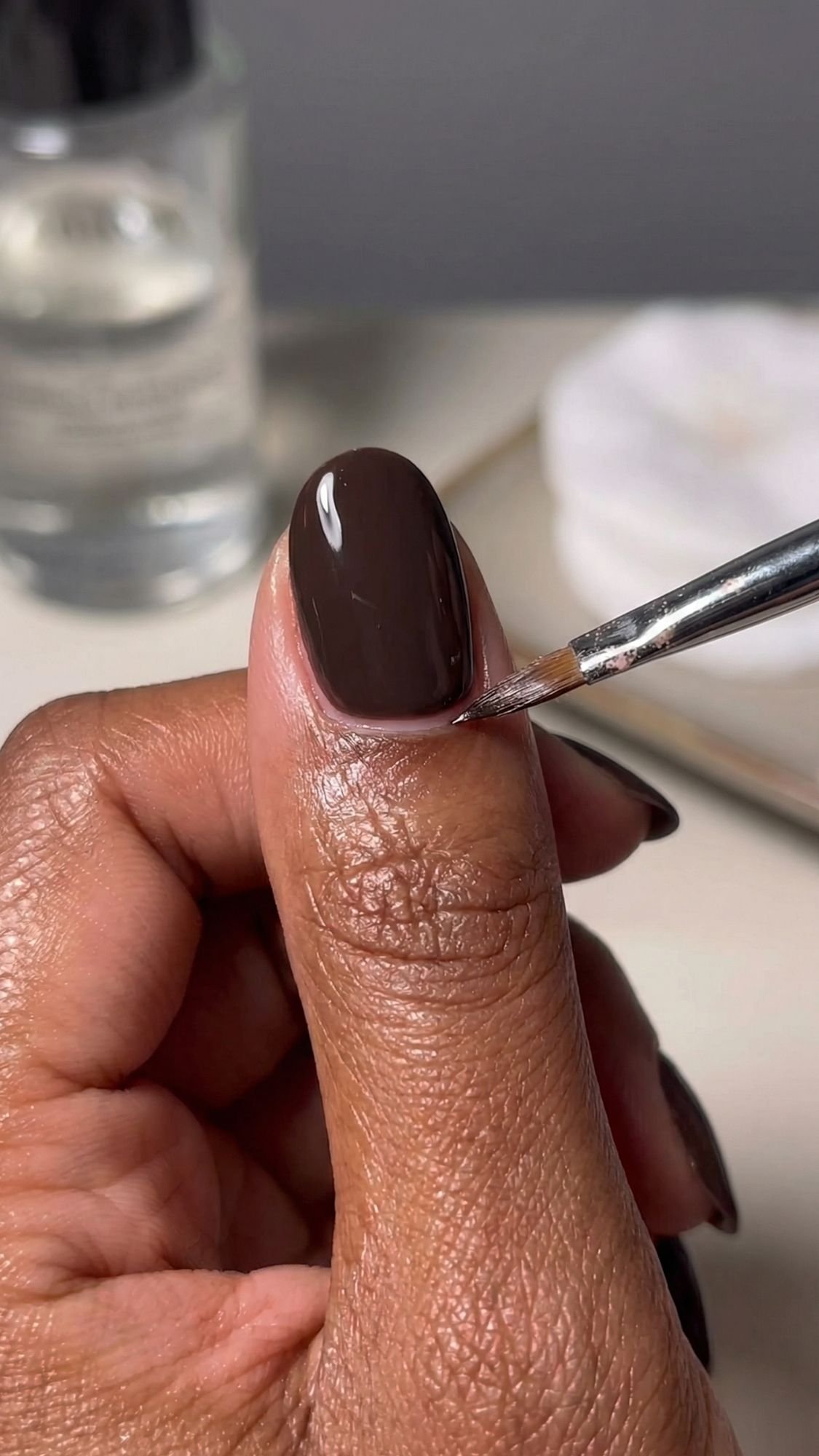A close-up of a hand with brown nail polish being applied to the thumb using a small brush—perfect inspiration for easy nail designs and creating simple nails at home. A bottle and white pad are blurred in the background.