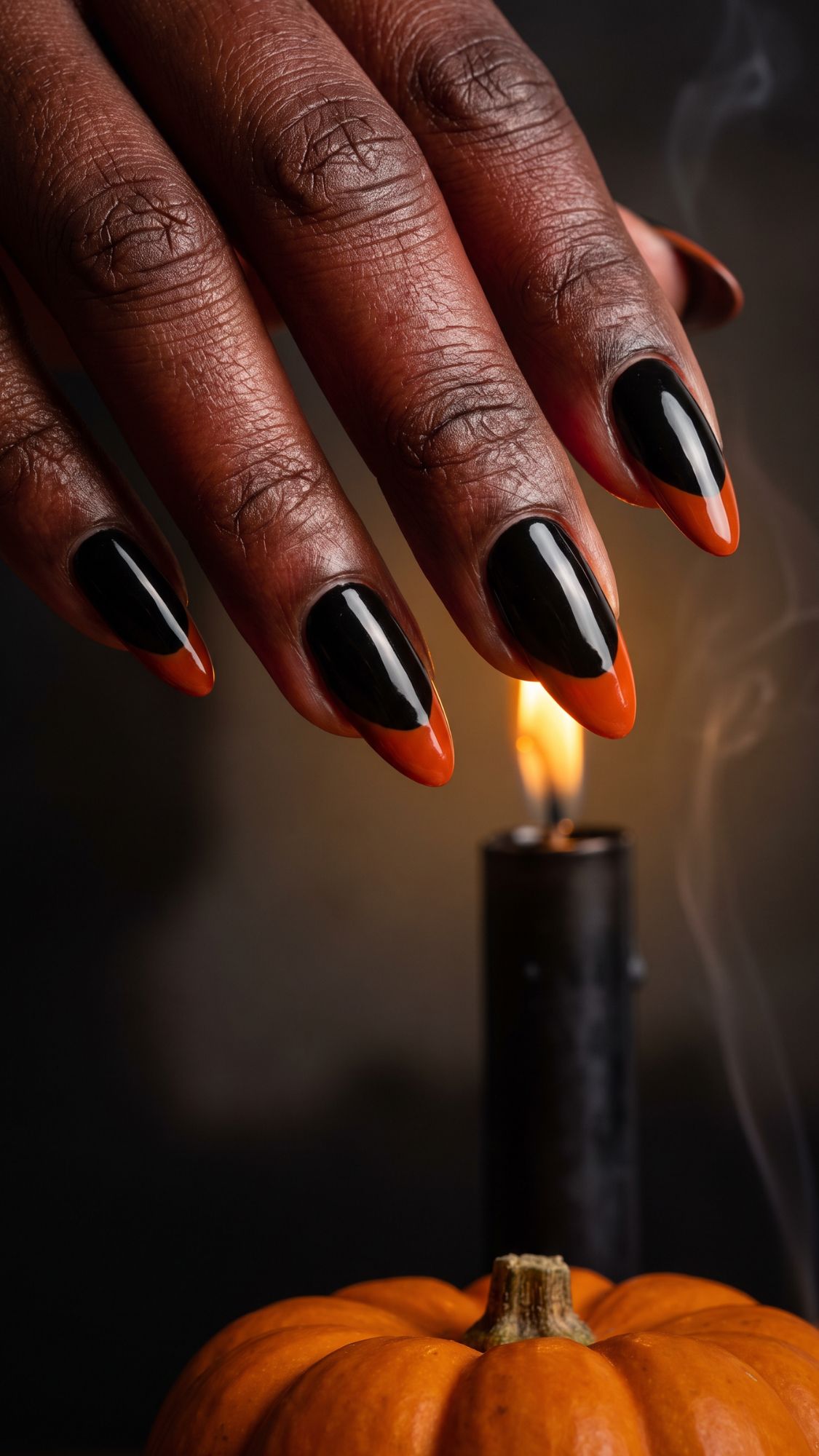 A hand with black and orange pointed nails hovers above a lit black candle. Showcasing glamorous Halloween Nail Designs, this scene is completed by a small orange pumpkin in the foreground, creating an irresistible Halloween-inspired vibe.