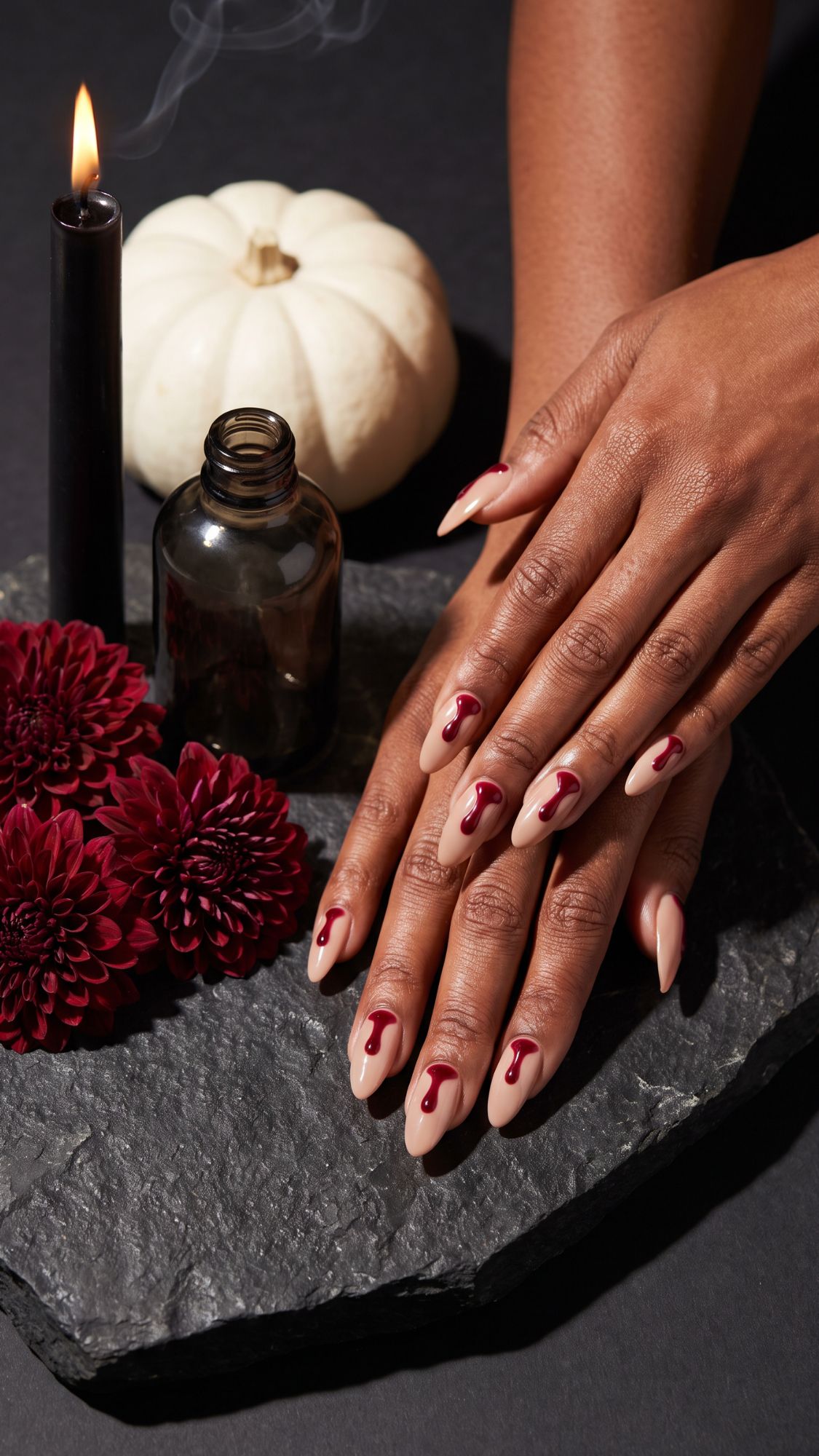 A person’s hands with nude nails featuring red drip designs rest on a black stone slab, showcasing glamorous Halloween nail designs. Nearby are a black candle, a dark glass bottle, dark red flowers, and a white pumpkin. The scene is moody and Halloween-themed—perfect inspiration for subtle Halloween nails with an elegant twist.