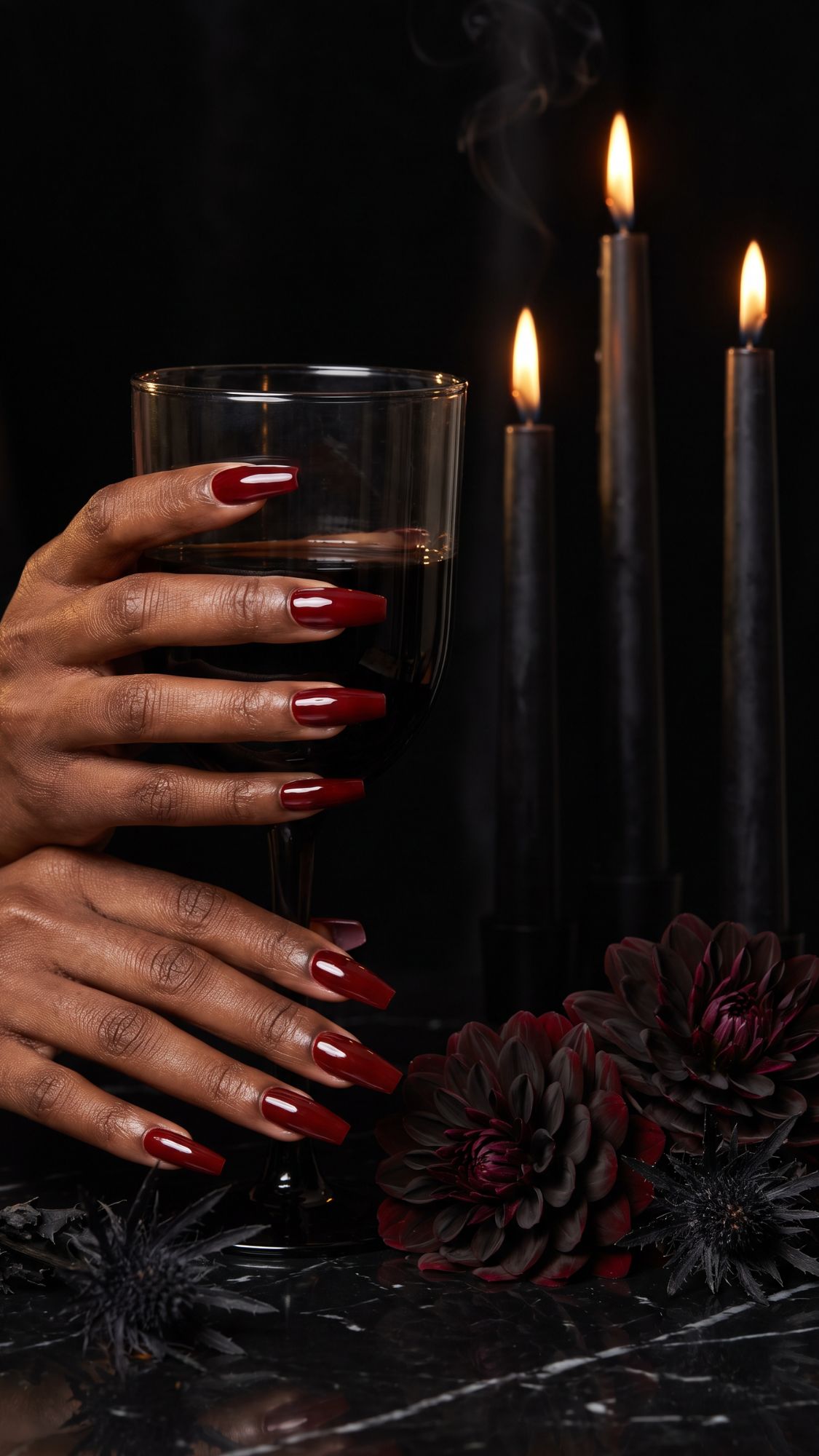 A hand with long, glossy red nails—perfect for glam Halloween nails—holds a wine glass. Three black candles burn in the background, next to dark dahlias on a marble surface. The scene is dramatic and moody.