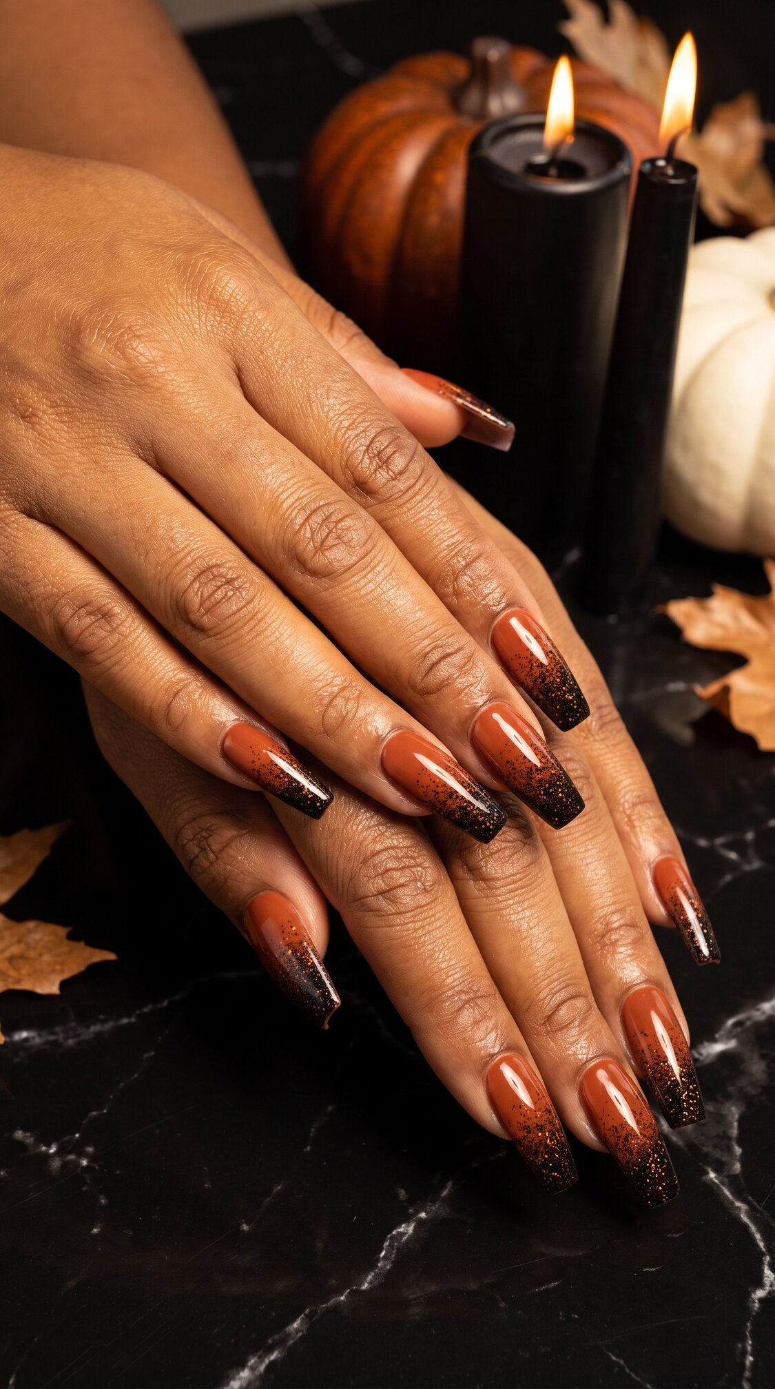 A person with long, brown and black glitter ombre halloween nails acrylic rests their hands on a marble surface. Black candles, pumpkins, and autumn leaves are in the background, creating a fall-themed setting.