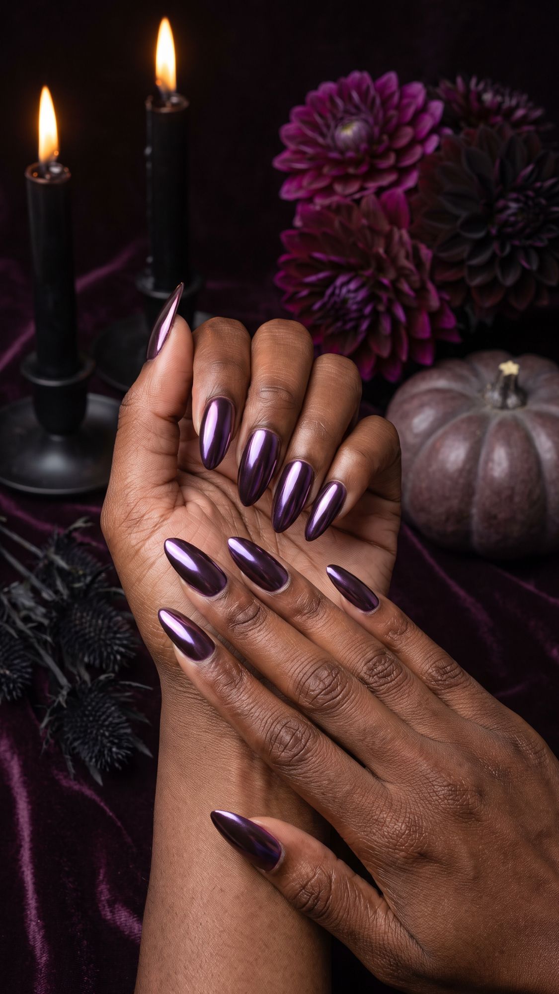 Hands with long, glossy, deep purple Glam Nails are shown against a dark background with two lit black candles, purple flowers, a velvet cloth, and a purple pumpkin, creating a gothic, elegant atmosphere perfect for Halloween Nails.