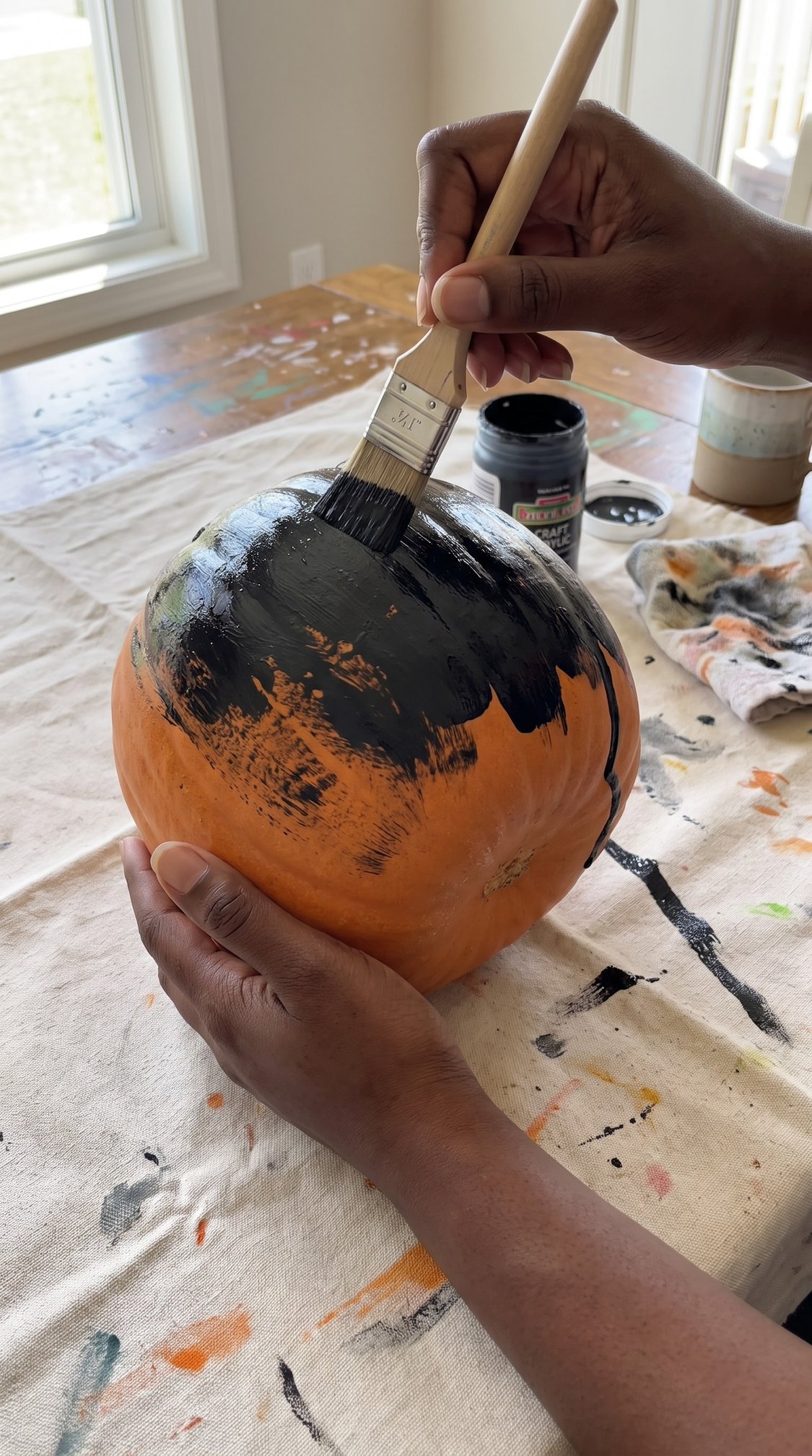 A person uses a paintbrush to paint the top of an orange pumpkin black. The pumpkin rests on a cloth-covered table with paint stains, and art supplies are visible in the background.