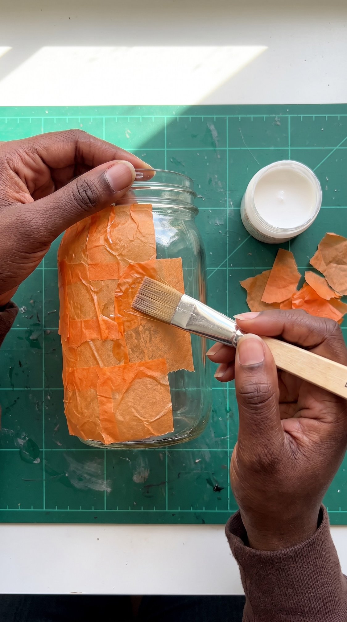 A person glues orange tissue paper pieces onto a glass jar using a paintbrush. A small jar of glue and more tissue paper are on a green cutting mat.