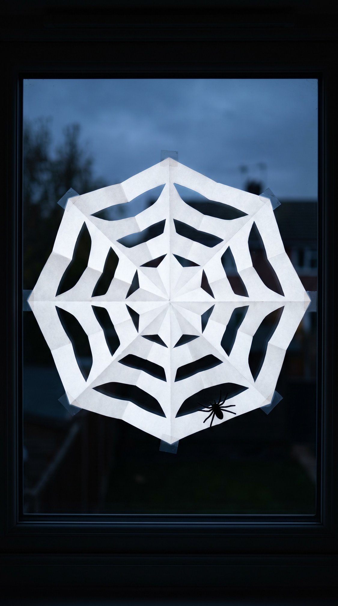 A paper snowflake cut into a spider web shape is taped to a window. A black paper spider is attached to the web. It is dusk outside.