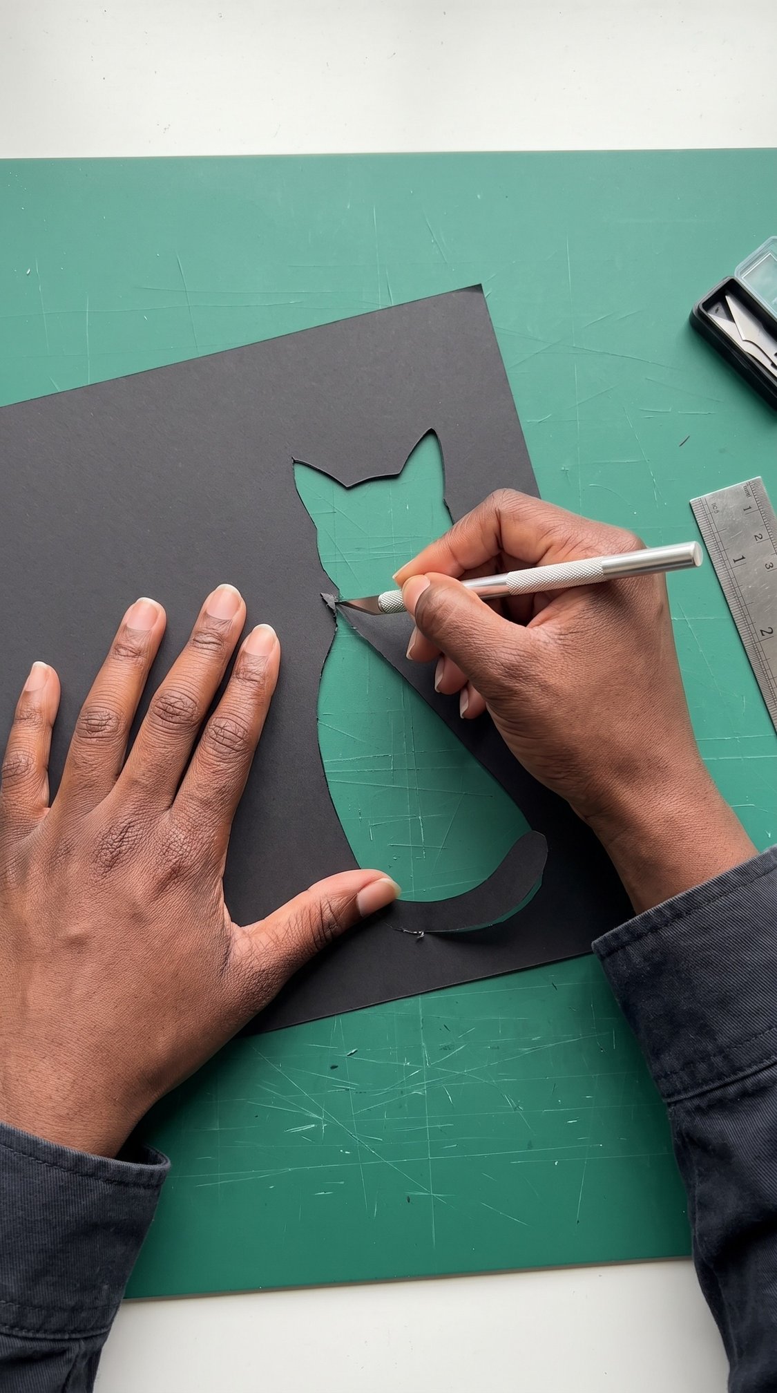 A person uses a craft knife to cut a cat silhouette from black paper on a green cutting mat. A metal ruler and a paper cutter are nearby.