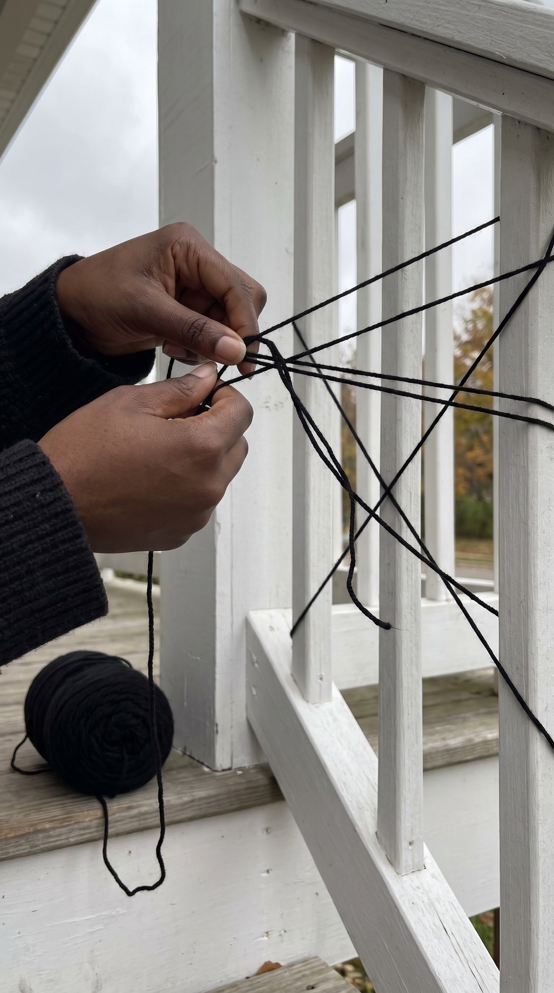 A persons hands string black yarn around white porch railings, creating a web-like pattern. A ball of yarn rests on the wooden floor nearby. The person is wearing a dark sweater.