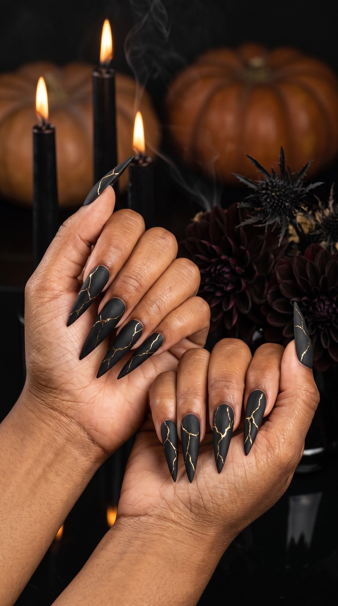 A pair of hands with long, pointed black halloween nails acrylic featuring gold crackle designs. In the background are lit black candles, dark flowers, and pumpkins, creating a spooky, Halloween-themed setting.