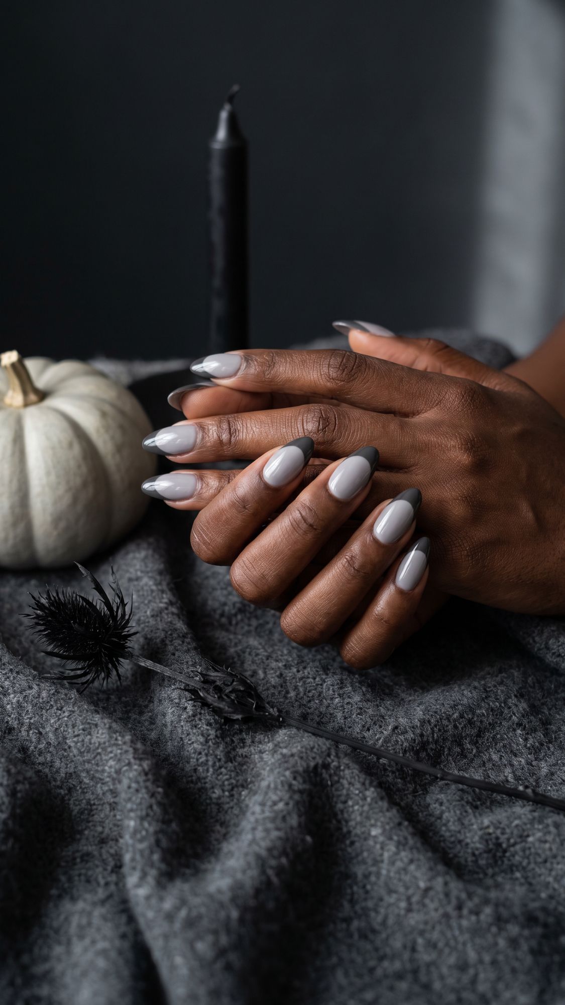 Hands with long, glossy gray nails rest on gray fabric. A white pumpkin, black candle, and black ornamental object are nearby, creating a moody, stylish autumn scene that perfectly showcases glam nail designs for Halloween.