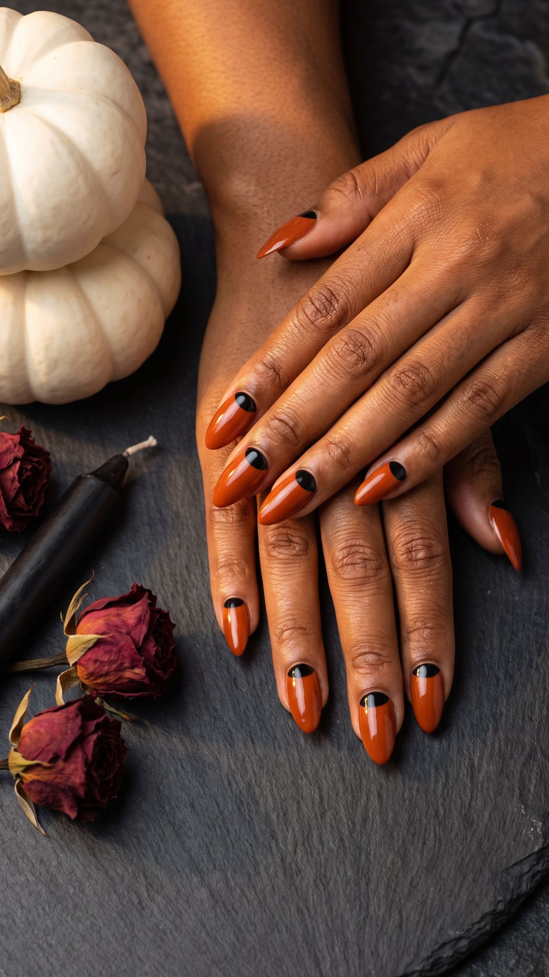Hands with long, orange and black geometric nail art rest on a dark surface near white pumpkins, dried red roses, and a black candle, creating a fall-themed, elegant setting that perfectly showcases chic Halloween nail designs.