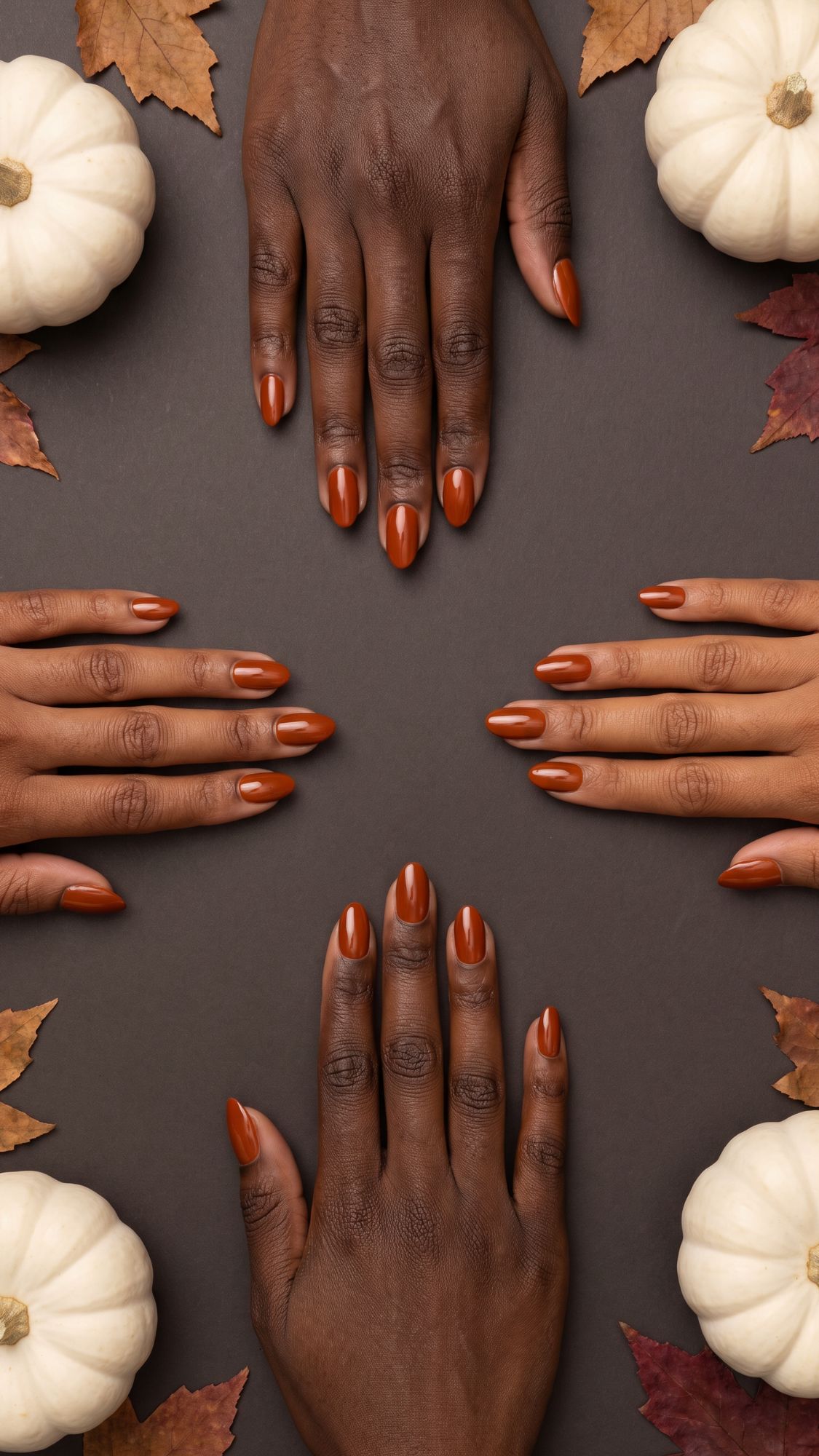 Four hands with brown skin and rust-colored nail polish are arranged on a dark background, surrounded by white pumpkins and autumn leaves, creating a fall-inspired composition that highlights subtle Halloween nails for a chic seasonal look.